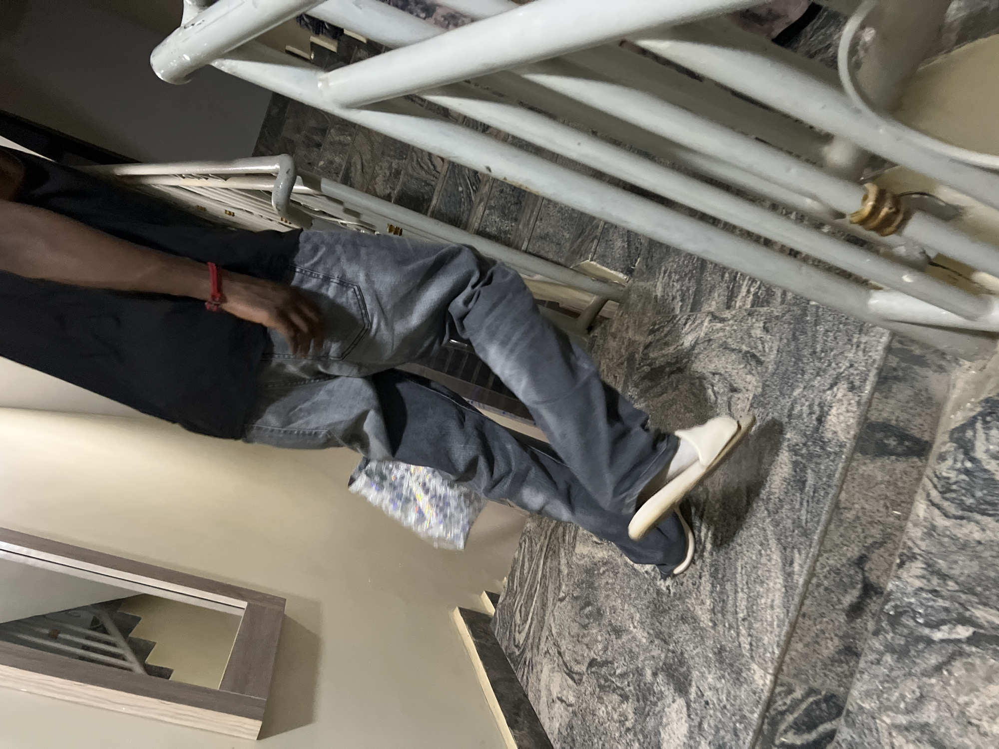 The image shows a person on a staircase, captured from a slightly elevated perspective. The individual, who has dark skin, is dressed in a dark long-sleeved top and dark grey jeans. A red bracelet adorns their right wrist. They wear white slip-on sandals on their feet. A small, shiny object, possibly a reflective bag or decorative item, is tucked into the back right pocket of their jeans.
The staircase itself is constructed from a dark grey and white mottled granite or marble. A prominent white metal railing, featuring several horizontal bars, runs alongside the stairs; some rust is visible on one of its connections. To the left, a rectangular mirror with a wooden frame is mounted on a light-colored wall, reflecting another section of a staircase.
The person appears to be standing or moving on the stairs, with their right foot positioned on a lower step. The scene is illuminated by artificial light, indicating an indoor setting, possibly during the evening or night. The overall environment suggests an interior space within a building in Gwarinpa, Nigeria. No text is visible, and the person's emotions cannot be discerned.