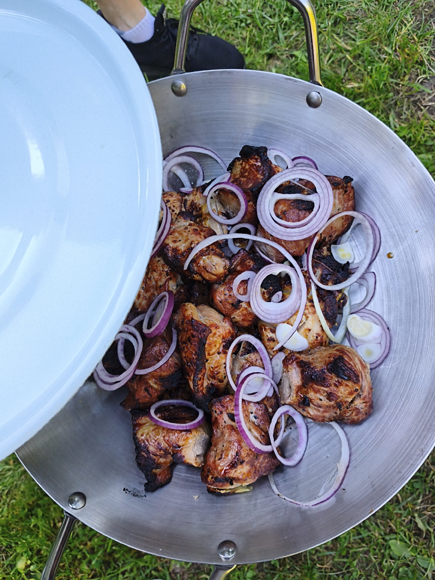 A large, round, metal grilling pan sits on the grass, filled with grilled meat and sliced red onions. The meat appears to be shashlik, with visible grill marks and some charred edges. The onions are thinly sliced rings. A large, white lid is partially covering the pan from the left. In the background, out of focus, a person is visible wearing a white sock and a black shoe, and green grass is present. The setting appears to be outdoors, likely in a yard or park. There is no specific indication of the city of Maardu, Estonia within the image itself.