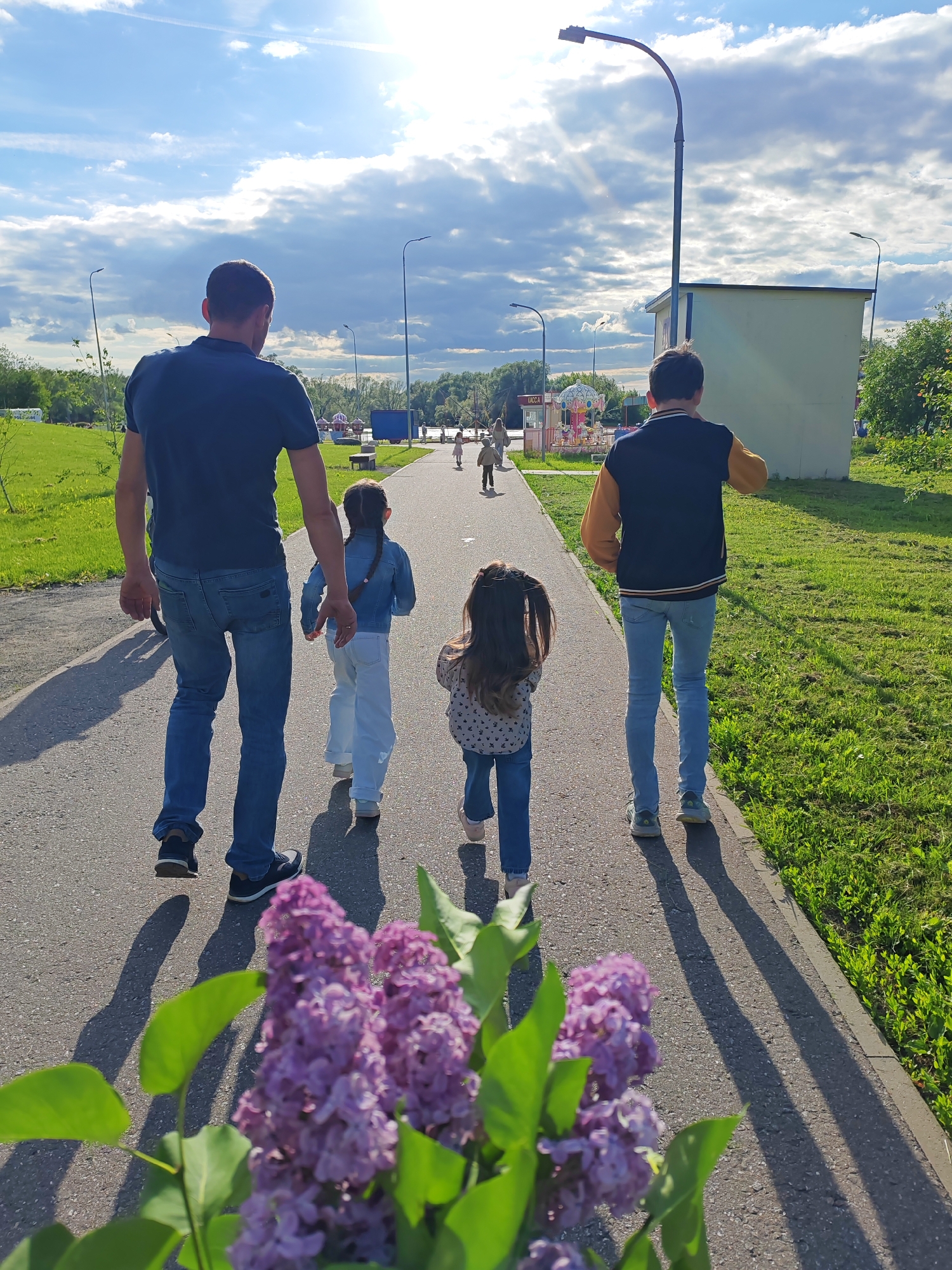 The image captures a family outing in Bronnitsy, Russia. A man, dressed in a dark blue t-shirt and jeans, walks with two young girls. Another person in a black and mustard yellow bomber jacket and jeans walks with them. In the foreground, a bouquet of purple lilacs adds a touch of color. The group is walking on a paved path that leads through a park with green grass on either side. In the background, there is a small building, street lamps, and a glimpse of a playground. The sky is partly cloudy, suggesting a pleasant day.