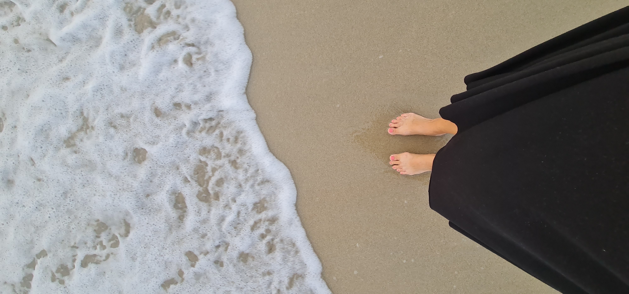 A top-down shot shows a person standing on a beach in Đà Nẵng, Vietnam. The person is wearing a long black dress that falls to their feet, which are visible against the tan sand. The toes are painted pink. To the left, white foamy waves wash onto the shore. The scene depicts the interaction between the person, the sand, and the ocean.
