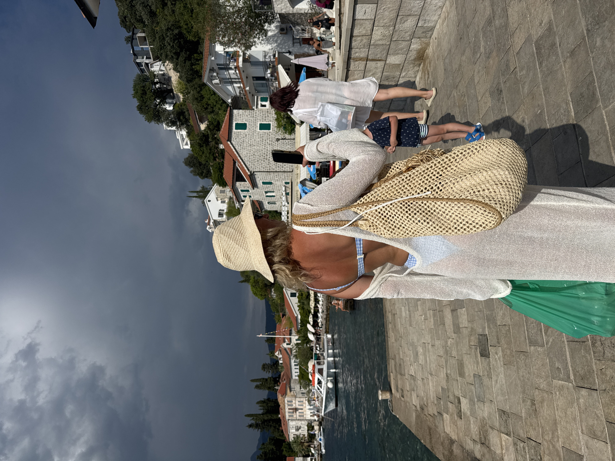 In the city of Unknown, Unknown, a woman wearing a straw hat and a sheer white cover-up over a blue and white checkered bikini stands on a stone pier, facing away from the camera. She holds a smartphone in her right hand, appearing to take a photo. A large, woven straw bag hangs from her left shoulder, and a bright green plastic bag is clutched in her left hand. In the background, the sea is calm, with several boats docked along the waterfront. Buildings with red-tiled roofs line the shore, and lush green trees and vegetation cover the hillside behind them. The sky above is a mix of bright sunlight and dark, ominous storm clouds. Further down the pier, another woman in a white cover-up walks away from the camera, accompanied by a young child in a dark blue shirt and striped shorts.