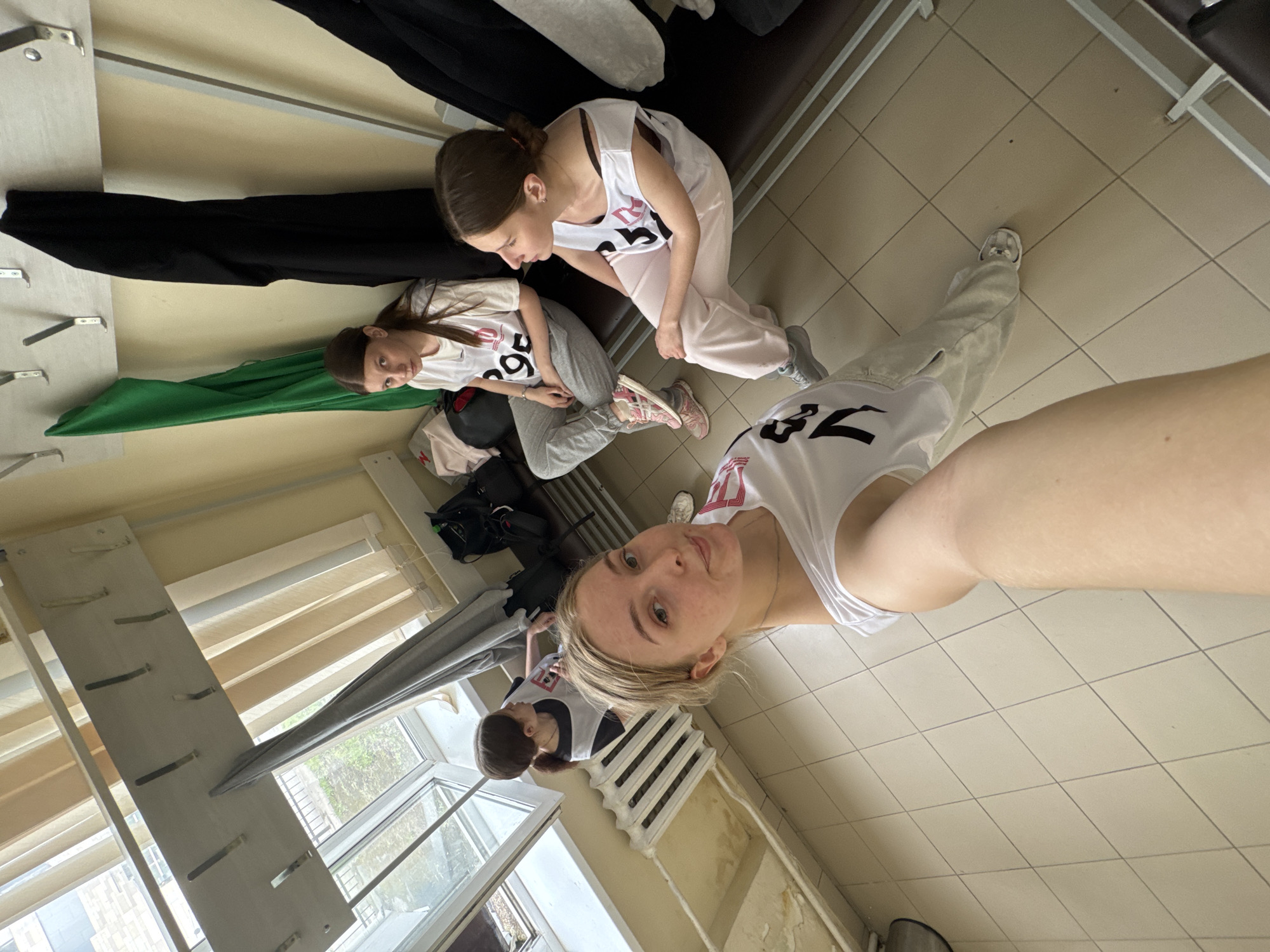 The image shows a group of three young women inside a room, possibly a dressing room or backstage area. The woman closest to the camera is taking a selfie, wearing a white tank top with the number "32" on it and light-colored sweatpants. She has blonde hair and is fair-skinned. Two other women are visible in the background, also wearing similar white tank tops with numbers on them. One has the number "396," and the other has no number visible. They are sitting or kneeling near a bench with clothing hanging above it. A rack or wall-mounted organizer is visible near a window. The room has tiled floors and appears to be indoors. The location and connection to the city of Perm, Russia, are not directly discernible from the visible contents of the image.
