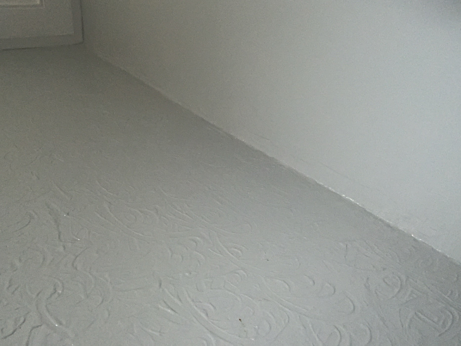 A corner of a room is shown, where the floor and two walls meet. The floor has a textured surface with a swirling, decorative pattern. The walls are smooth and painted a similar light color as the floor. The lighting is somewhat dim and diffused, creating a soft contrast between the surfaces. Part of a doorframe is visible in the upper left corner.