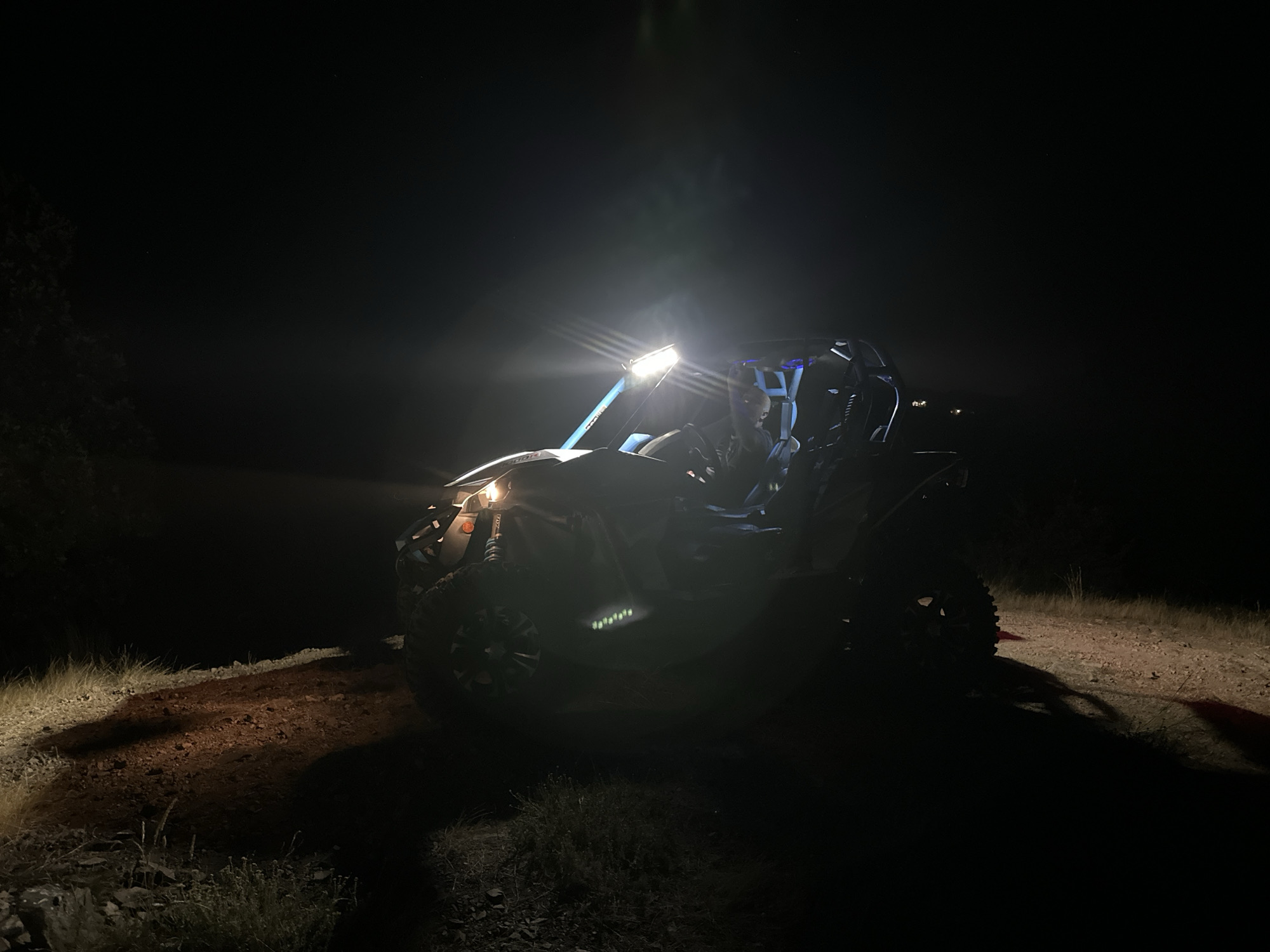 A dark, low-angle, wide shot shows a side-by-side vehicle illuminated by its headlights and a roof-mounted light bar at night. The vehicle is on a dirt and gravel road. The background is dark and indistinct, with some faint illumination in the distance. The scene is devoid of any specific identifiable landmarks that would indicate the location in Momin Prohod, Bulgaria. No people are clearly visible within the vehicle. The lighting casts long shadows across the road and surrounding terrain.
