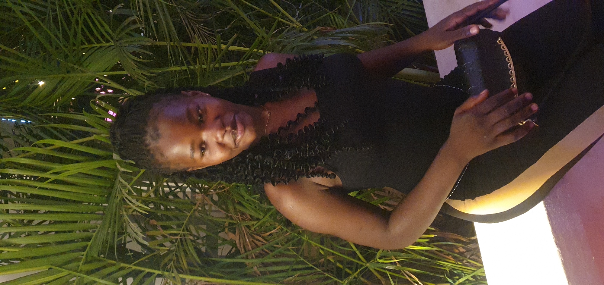 In Bar Jirgi Summa, Nigeria, a young Black woman with braided hair is seated against a backdrop of lush green palm leaves. She is wearing a black sleeveless top and black pants with a distinctive yellow stripe down the side. In her hands, she holds a small black clutch purse adorned with gold accents. The lighting suggests it is evening or night, with artificial lights illuminating the scene from the front and possibly from behind the foliage, creating a warm glow. She is looking towards her left, with a subtle smile on her face.
