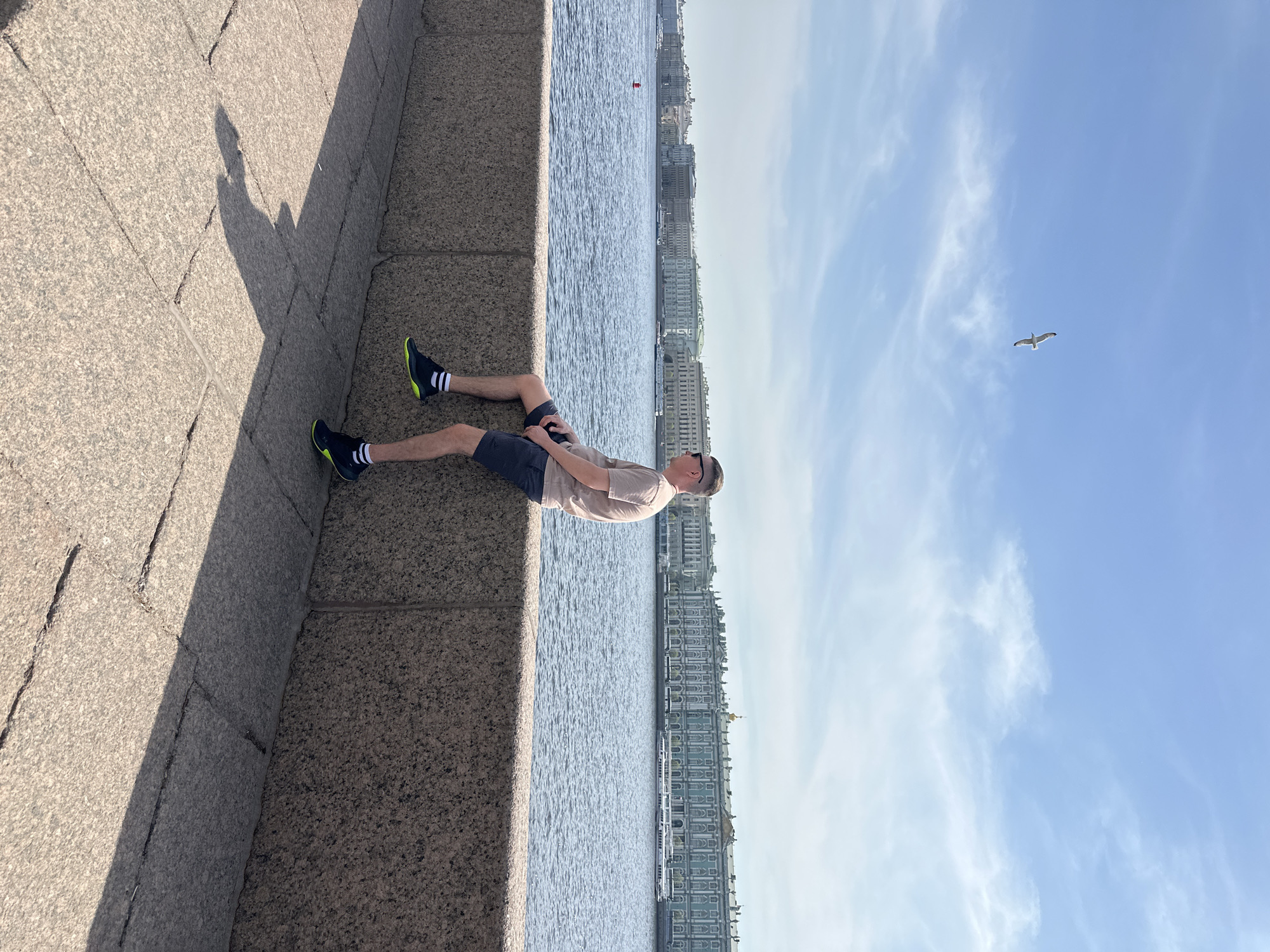 A man is sitting on a granite embankment in Saint Petersburg, Russia. He is wearing a tan shirt, dark shorts, black and neon green shoes, and white socks with black stripes. He has dark sunglasses and a light complexion. He appears to be looking at his phone. The Neva River is visible in the background, along with several historical buildings lining the embankment. The sky is blue with light clouds, and a single bird is flying in the upper right corner of the image. The granite embankment casts a shadow on the stone pavement where the man is sitting.