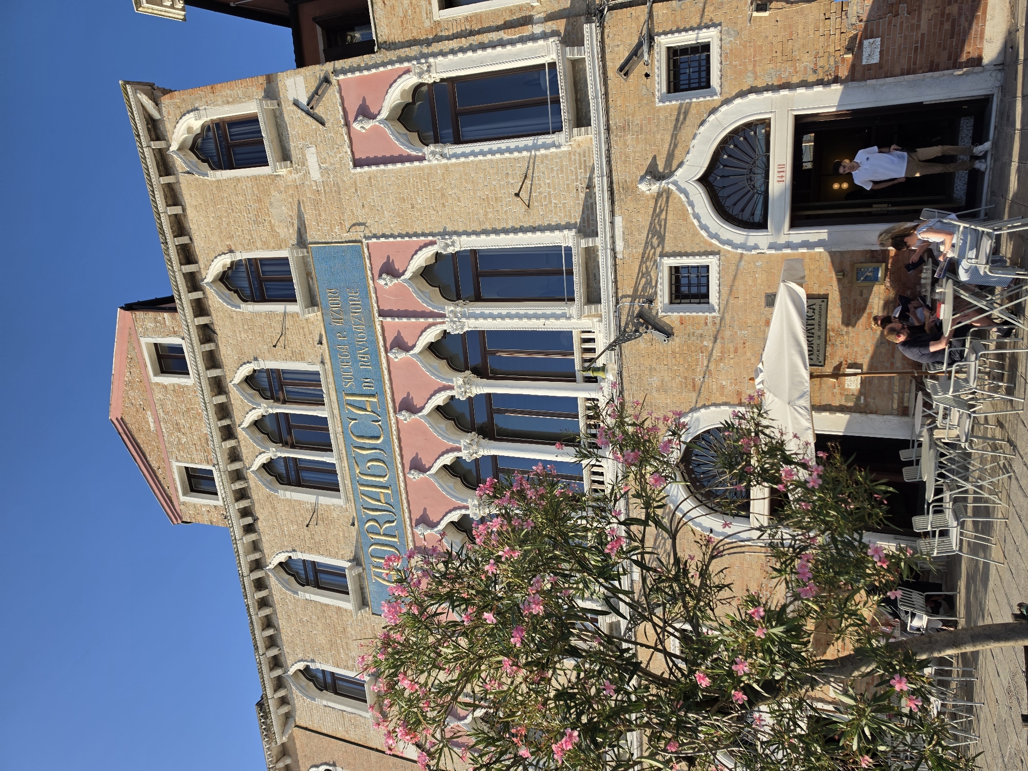 The image captures the facade of a historical building in Venice, Italy, likely near a canal, as suggested by the architecture and outdoor seating area. The building features characteristic Venetian Gothic architectural elements, including pointed arches in the windows and decorative stonework. A prominent blue sign with gold lettering reads "ADRIATICA SOCIETA P. AZIONI DI NAVIGAZIONE," indicating it was once a shipping company. In the foreground, a table and chairs are set up outside, suggesting a cafe or restaurant. A tree with pink flowers is positioned to the left of the building. Several people are seated at the tables, engaged in conversation and reading. A young man in a white polo shirt and khaki pants stands in the doorway of the building. The sky is clear and blue, indicating a sunny day.