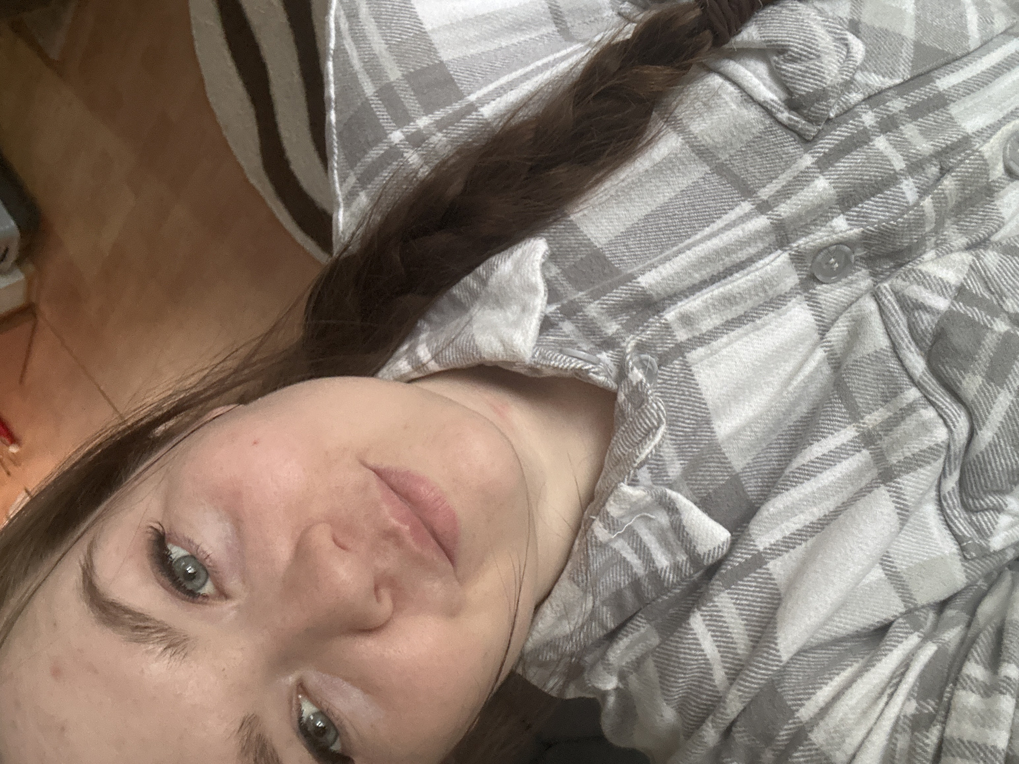 A selfie of a woman with fair skin and light blue eyes, wearing a gray and white plaid button-down shirt. Her brown hair is braided. The image is taken at an angle, with the woman's face slightly tilted towards the camera. The background is out of focus, but it appears to be indoors, with part of a striped rug visible in the upper left corner. There is no definitive evidence to suggest the location is in Chaikovsky, Russia.