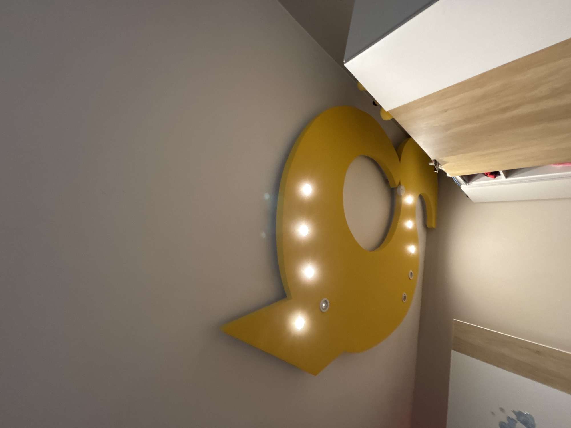 A sideways view reveals a yellow, illuminated letter "Q" mounted on a light gray wall. The letter features multiple light bulbs, with some appearing brighter due to proximity or reflection. Above the "Q" is a portion of a built-in structure with a white top and a wood-grained base, suggesting it could be a cabinet or shelf unit. Further into the scene is a glimpse of what appears to be the interior of a room, including sections of a wall and possibly a frame or piece of art. Based on the style, it could be a modern room, maybe a child’s bedroom, in a location that is in Sofia, Bulgaria.