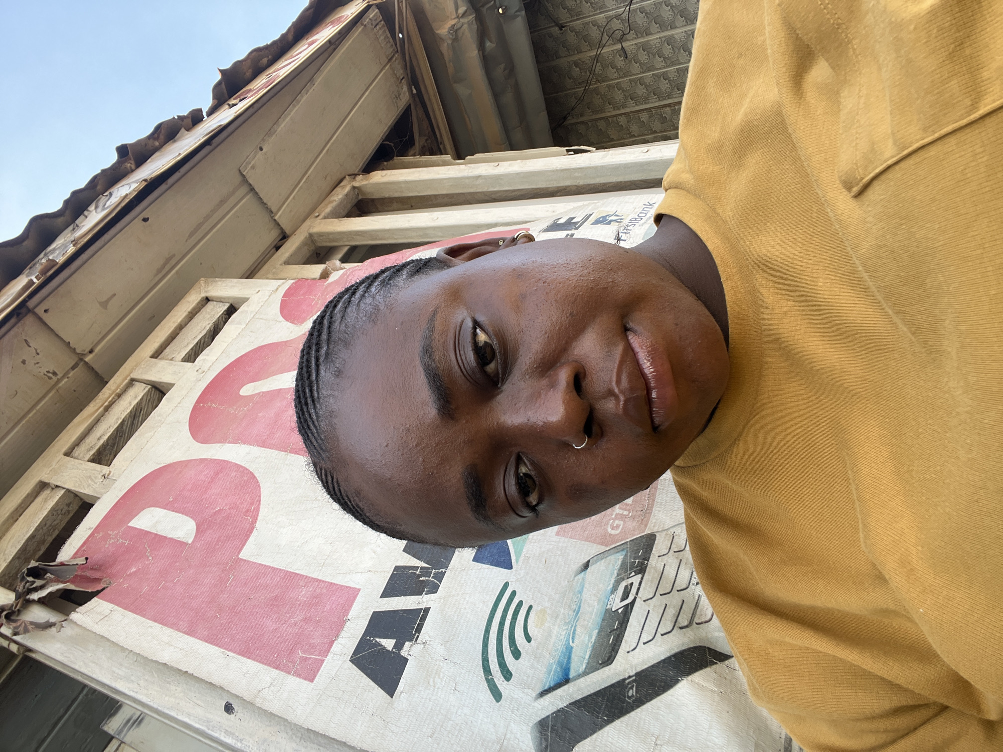 A selfie is taken by a young Black woman with braided hair and a nose ring, wearing a mustard yellow t-shirt. She is looking directly at the camera with a neutral expression. The background appears to be an outdoor setting, possibly in a city. A large, faded banner with red lettering is visible behind her, with parts of the letters "P" and "A" discernable. Below the banner, there is an image of a mobile phone and what appears to be some form of signage that includes the words "FirstBank" and "GT". The overall environment suggests an urban setting, possibly a street or a market area, with signs of wear and tear on the structures. The lighting indicates it is daytime. The specific location in Abuja, Nigeria is not discernible from the image alone.