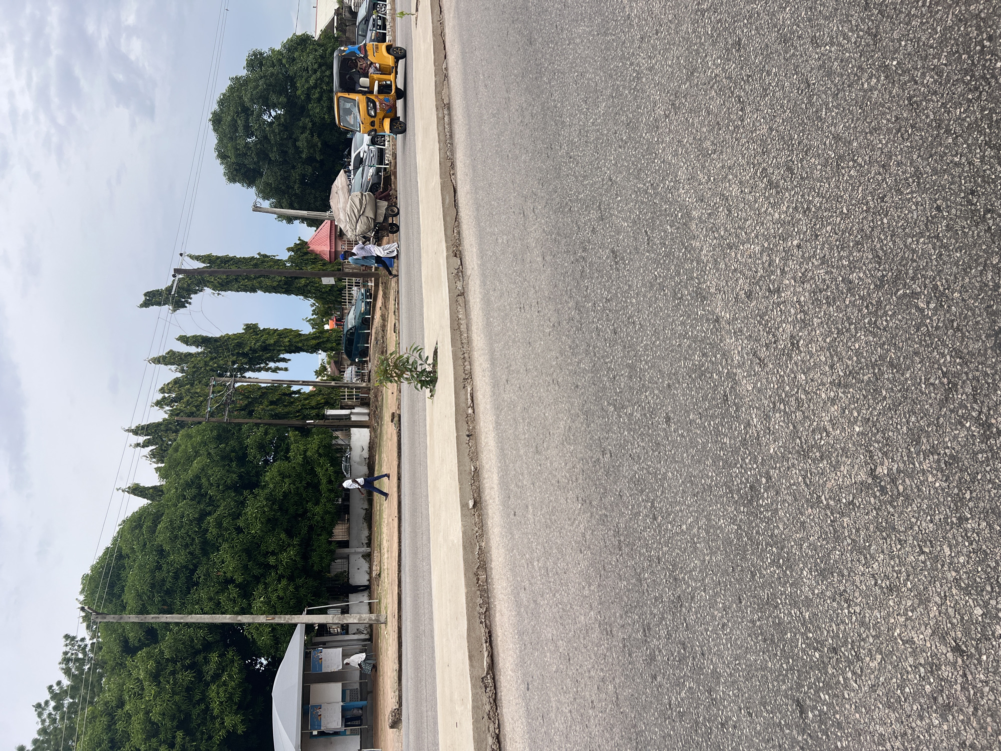 A street scene in Jalingo, Nigeria, features a paved road on the right, with a sidewalk bordering a grassy area on the left. Several vehicles are parked along the side of the road, including a yellow auto-rickshaw, other cars, and a cart loaded with goods covered by a tarp. Two people are visible on the sidewalk: one appears to be walking, while another is seated. The background shows lush green trees and utility poles with overhead wires. A few buildings with signs are also present in the distance. The sky is overcast, suggesting a cloudy day. The image is taken from a slightly elevated perspective, looking down the street. No discernible text is visible in the image. The overall impression is a typical day on a bustling street in a Nigerian town.