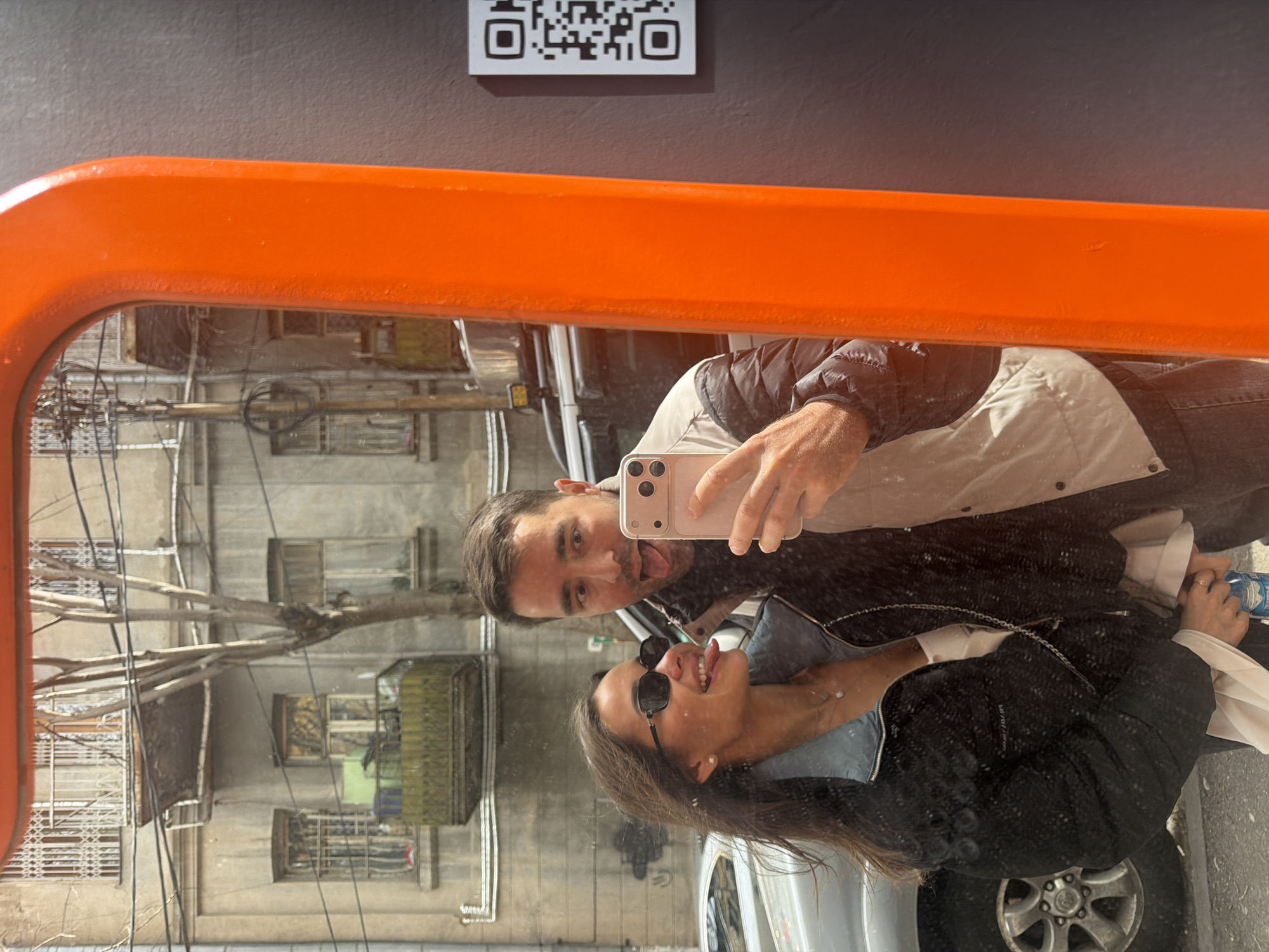 Reflection of a man and a woman taking a selfie in an orange-framed mirror. The QR code above the mirror suggests a public location. The background reveals a street scene with older buildings, trees, and utility lines characteristic of Tbilisi, Georgia. A car is visible in the lower right corner of the reflection. The woman wears sunglasses and a black jacket. The man holds the phone, sticking out his tongue.