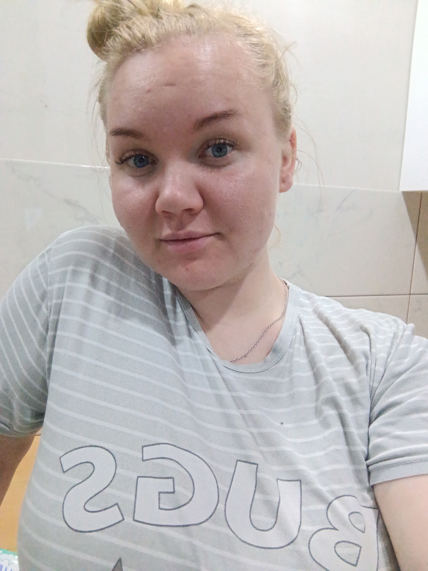 A fair-skinned woman with blonde hair in a bun is centered in the frame, wearing a gray and white striped t-shirt with the word "BUGS" printed on it. She is wearing a thin silver chain around her neck. The background is a neutral-toned wall, with possible tile or marble patterning in the upper left corner. The lighting is even, suggesting an indoor environment. There's no verifiable evidence in the image to place the location in the city of Фоки, Russia.