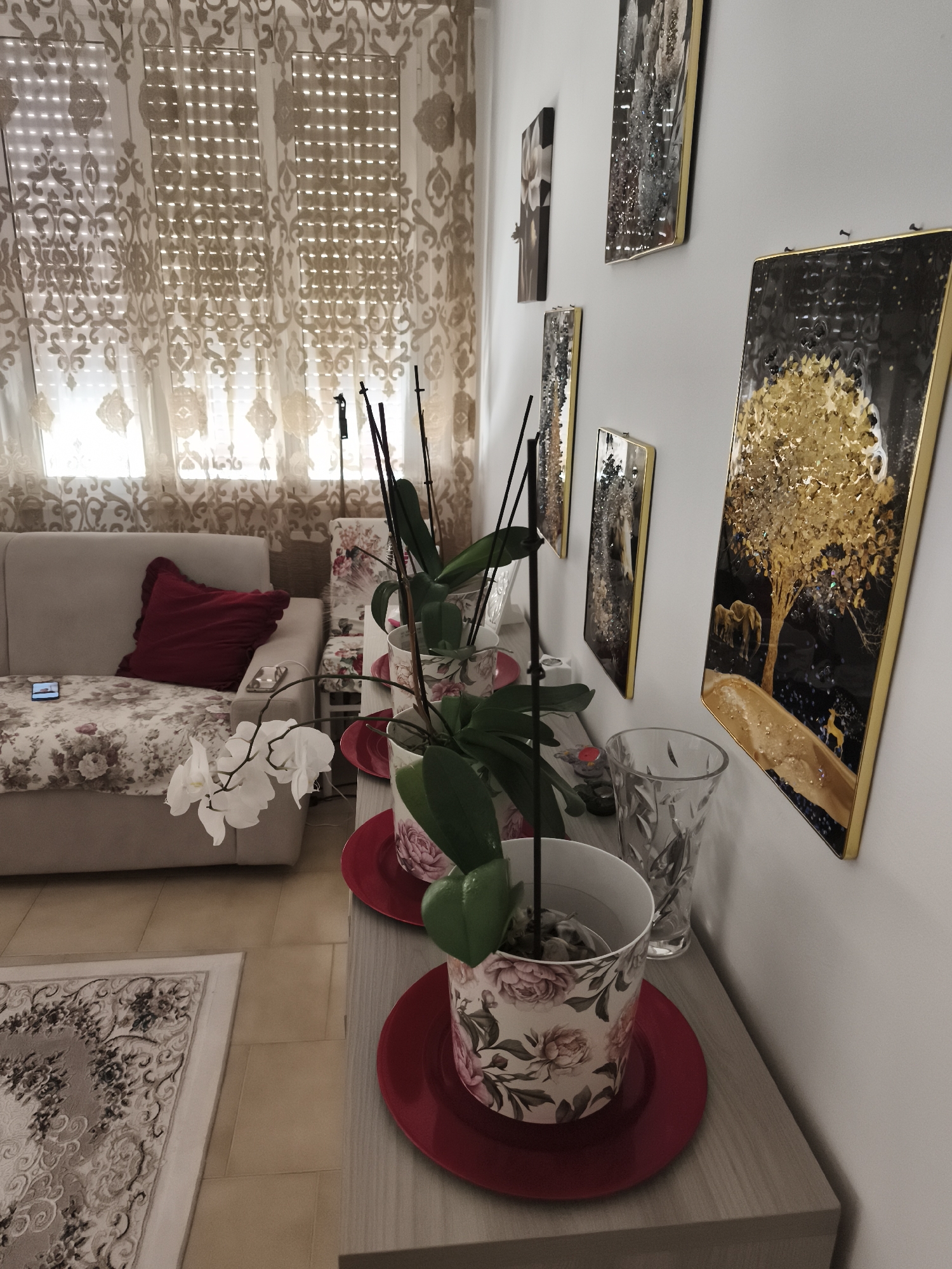 The image depicts an indoor scene, possibly a living room, with a focus on decorative items and furniture. In the foreground, several potted plants, identified as orchids by their white blooms, are arranged on a light-colored wooden shelf. The pots are adorned with a floral pattern featuring roses. Red saucers are placed beneath the pots. To the left of the plants, a sofa is visible, upholstered in a light-colored fabric with a floral pattern. A red throw pillow rests on the sofa. A smartphone is placed on the sofa's armrest.
In the background, large windows are covered by sheer, patterned curtains. On the wall to the right, a collection of framed artwork is displayed. These artworks appear to be modern and abstract, with a prominent use of gold and black colors, featuring imagery such as a golden tree with elephants and a deer. A clear glass vase is situated on the shelf next to the potted plants.
The floor is tiled in a light beige color and partially covered by an ornate rug with floral motifs. The overall lighting suggests daytime, with natural light filtering through the curtains.
The prompt specifically asks to include the location in the city of Livorno, Italy. However, there is no information within the image or any accompanying data that allows for the determination of a specific city or location. Therefore, the location in Livorno, Italy cannot be provided.