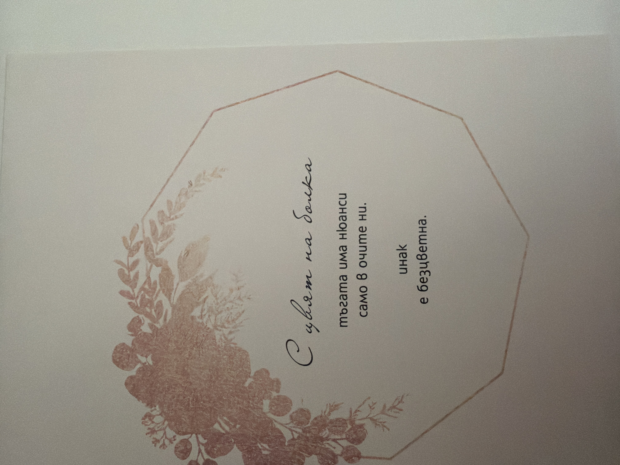 The image shows a close-up of a card with a floral design in shades of pink and a thin, geometric outline. Text is printed in dark grey font. The text reads: "С цвет на болка / тъгата има нюанси / само в очите ни. / инак / е безцветна." This translates to: "With the color of pain / sadness has nuances / only in our eyes. / otherwise / it is colorless." There is no indication of a specific location in Pazardzhik, Bulgaria, or any people or actions.