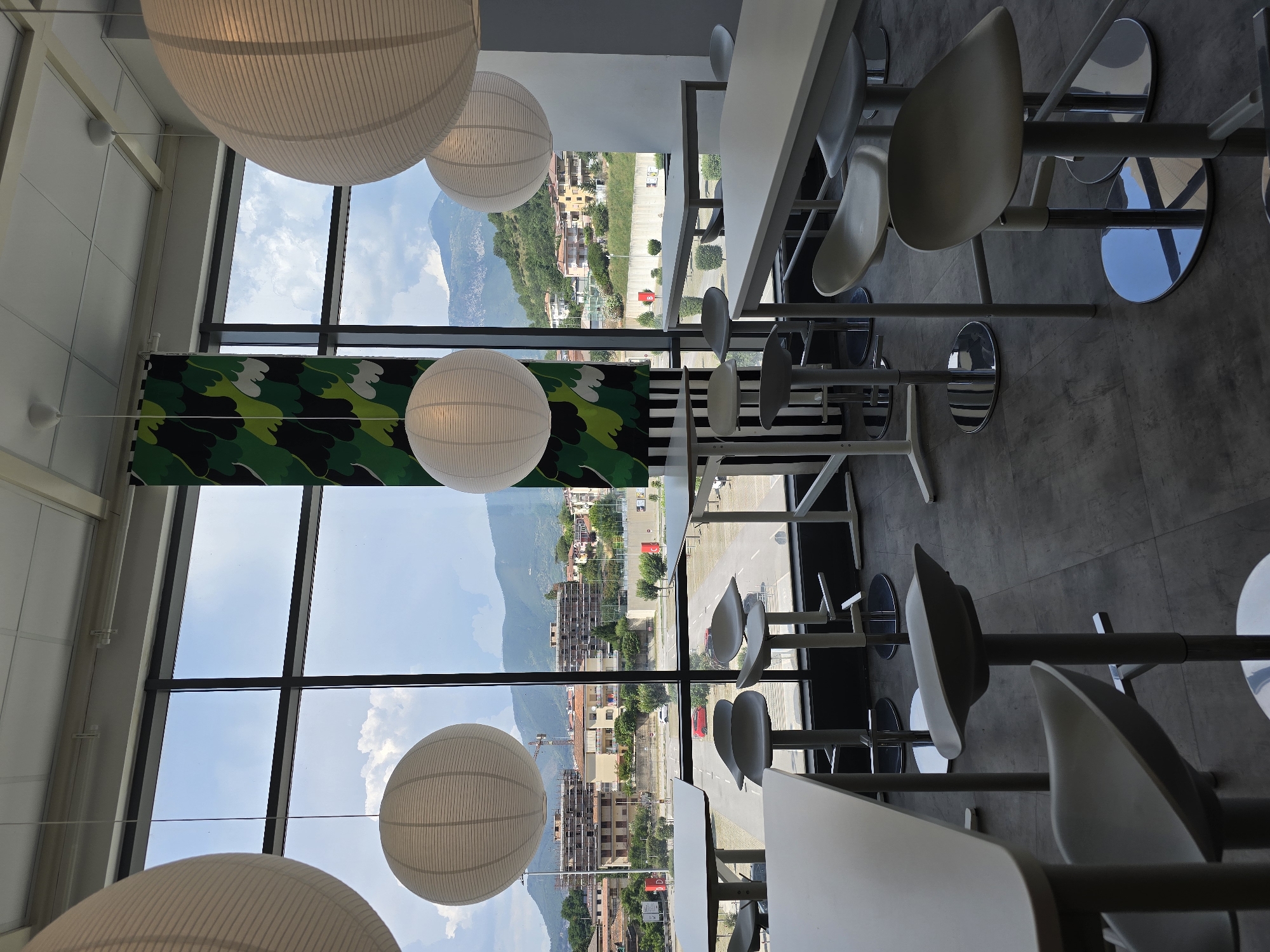 The image depicts the interior of a bright, modern cafe or restaurant, likely part of a chain or standardized establishment, given the consistent design elements. Large windows dominate the background, offering a panoramic view of a moderately sized Italian town, specifically identified as Baronissi, Italy, nestled at the foot of rolling green hills. The architecture visible outside suggests a mix of residential buildings, some appearing under construction with scaffolding, and commercial structures. The sky is partly cloudy.
Inside, the space is furnished with white, minimalist tables and high stools, many of which are also white or light grey, with a silver chrome base. Several large, spherical, white paper lanterns hang from the ceiling, providing soft illumination. A tall, narrow curtain with a black and green leaf-like pattern hangs vertically in front of one of the windows, adding a touch of decorative color. The flooring is a dark grey tile. The overall impression is one of a clean, contemporary dining or waiting area with a view. No people are visible in the scene.