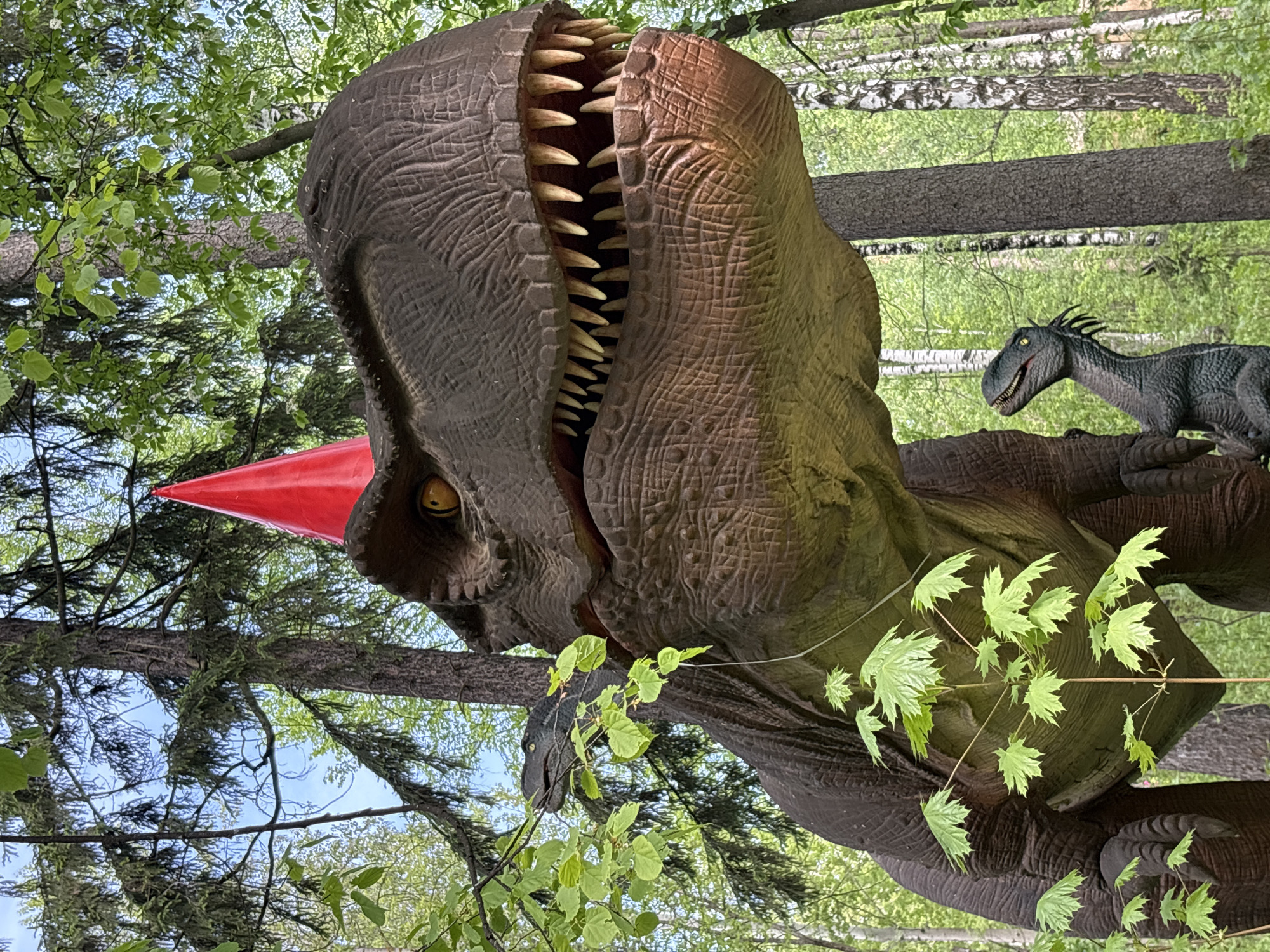 This outdoor photo depicts a large, brown, animatronic Tyrannosaurus Rex (T-Rex) dinosaur with an open mouth full of teeth. Attached to the T-Rex is a red cone at its head. Behind the T-Rex, there is a smaller animatronic dinosaur. The scene is set in a wooded area with trees and green foliage. The location is Izhevsk, Russia.