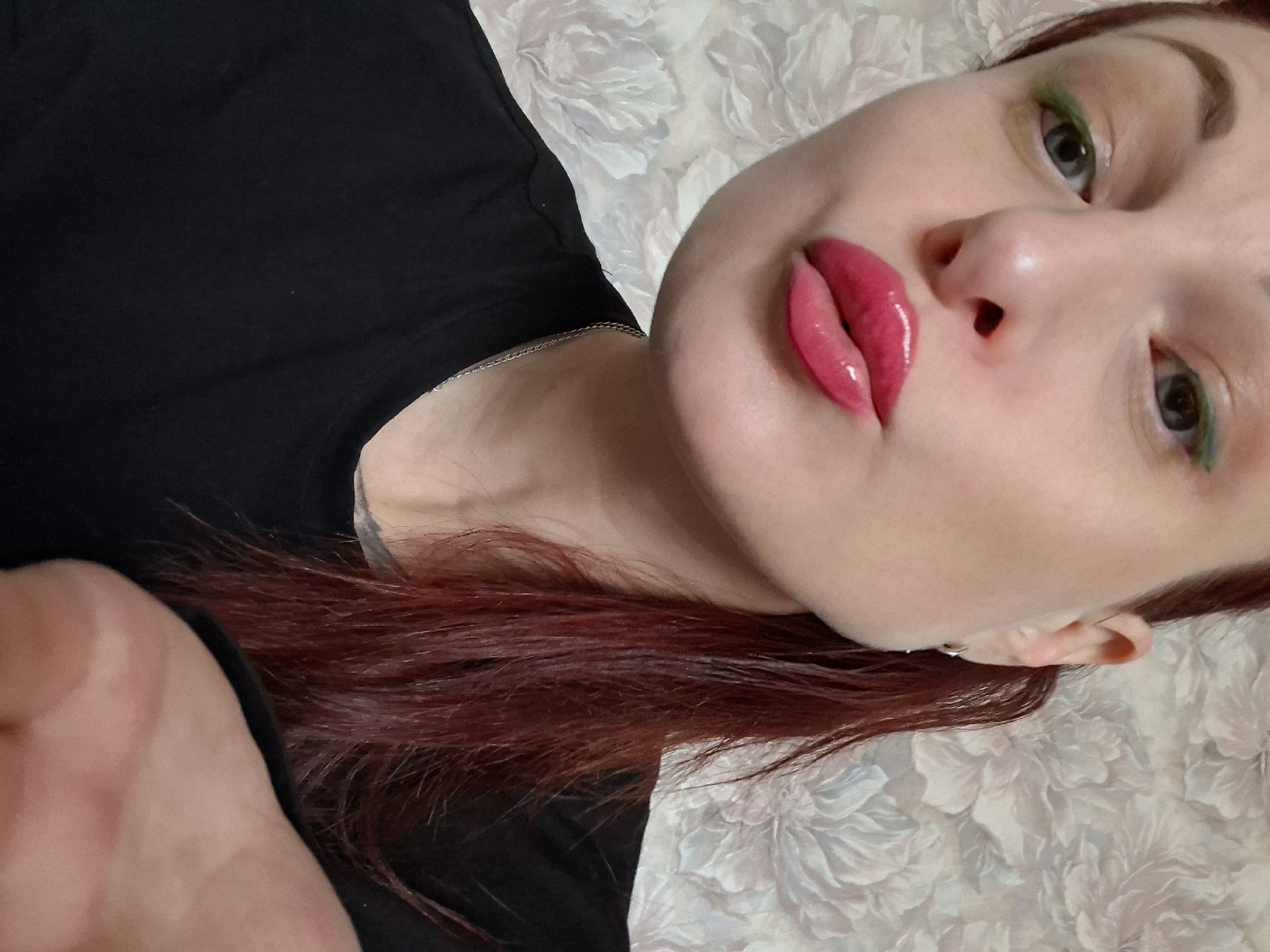 A selfie of a woman with red hair and bright pink lips. She is wearing a black shirt. Her eyes are partially lined with green eyeliner. She is lying on a floral-patterned surface. The location is Забалоцкі сельскі Савет, Belarus.