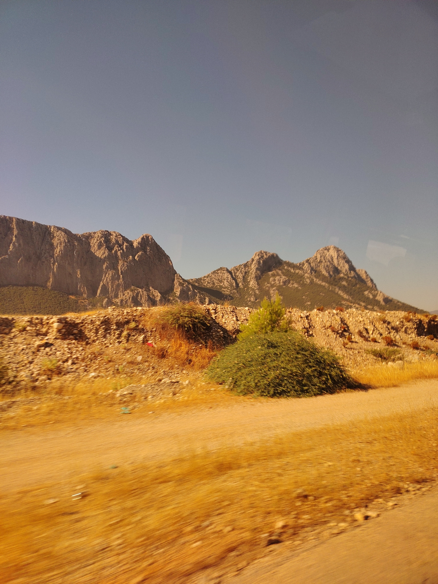 The image captures a landscape scene with prominent mountains in the background under a clear blue sky. In the foreground, a dry, rocky terrain is visible with sparse vegetation, including a dense green bush and some dry grasses. A dusty path or road runs through the scene. The mountains appear rugged and mountainous, with some vegetation clinging to their slopes. The overall impression is of a natural, somewhat arid environment. This scene is likely located in the region of Konyaaltı, Turkey, known for its natural beauty and mountainous terrain.