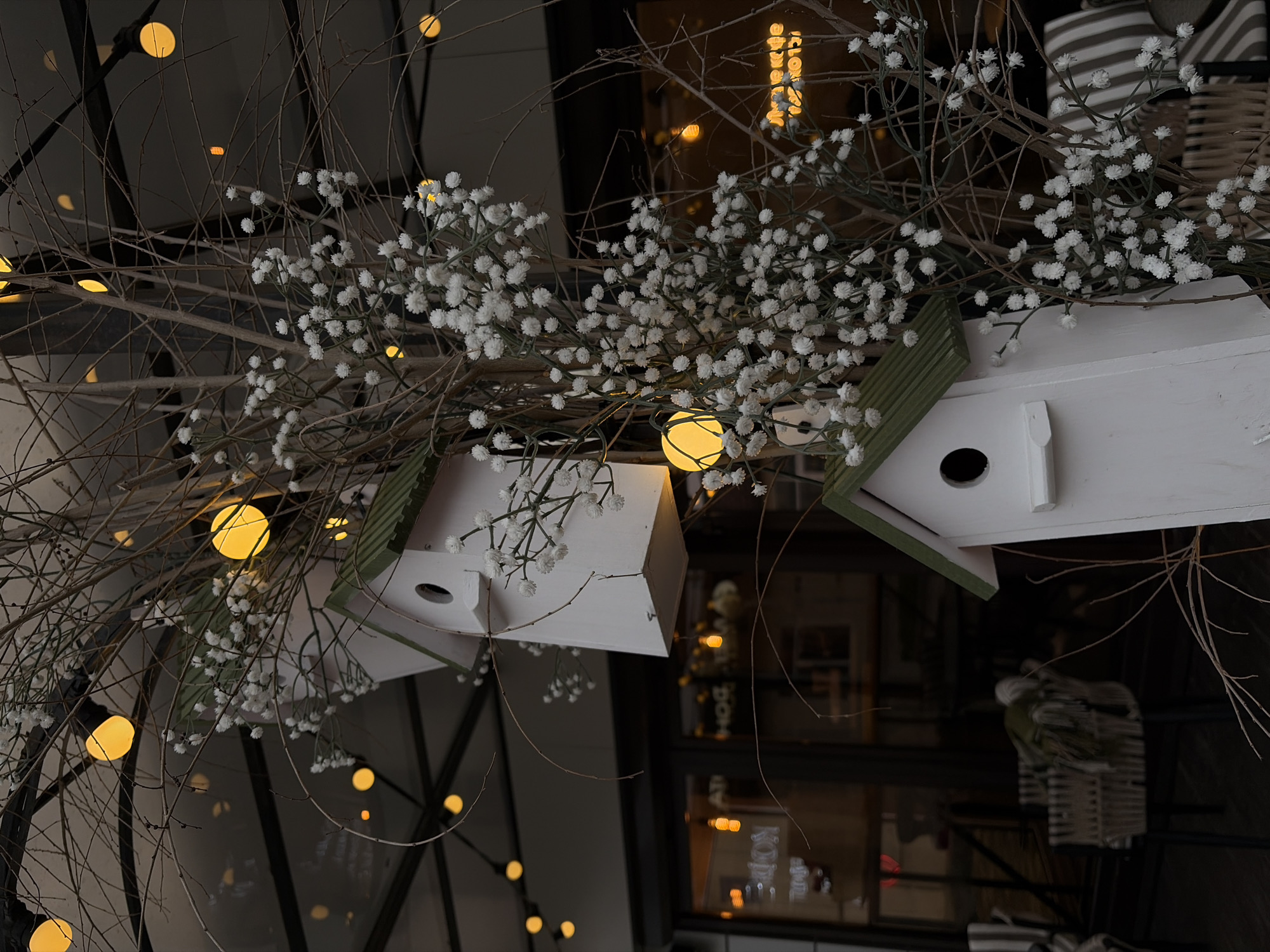 This upward-angled close-up photograph captures a decorative indoor display, likely in a cafe or public establishment in Novopokrovskaya, Russia. The main subjects are two white wooden birdhouses with green roofs, positioned at an angle, integrated into a dense arrangement of bare, dark tree branches.
Intertwined with the branches are numerous clusters of small, white artificial flowers, resembling baby's breath. Scattered throughout the branches are several warm, glowing yellow spherical lights, contributing to a cozy and inviting atmosphere, suggesting the image was taken during the evening or night.
The background is dark, featuring reflections from what appears to be a window or glass partition. Within these reflections, parts of an interior are visible, along with reversed text. The Russian word "КОФЕ" (KOFE), meaning "COFFEE," is clearly discernible, reinforcing the setting as a cafe. Additionally, elements like woven baskets with striped patterns are visible in the background, contributing to the rustic and decorative aesthetic of the scene. The overall impression is one of a charming, nature-inspired, and warmly lit interior design.