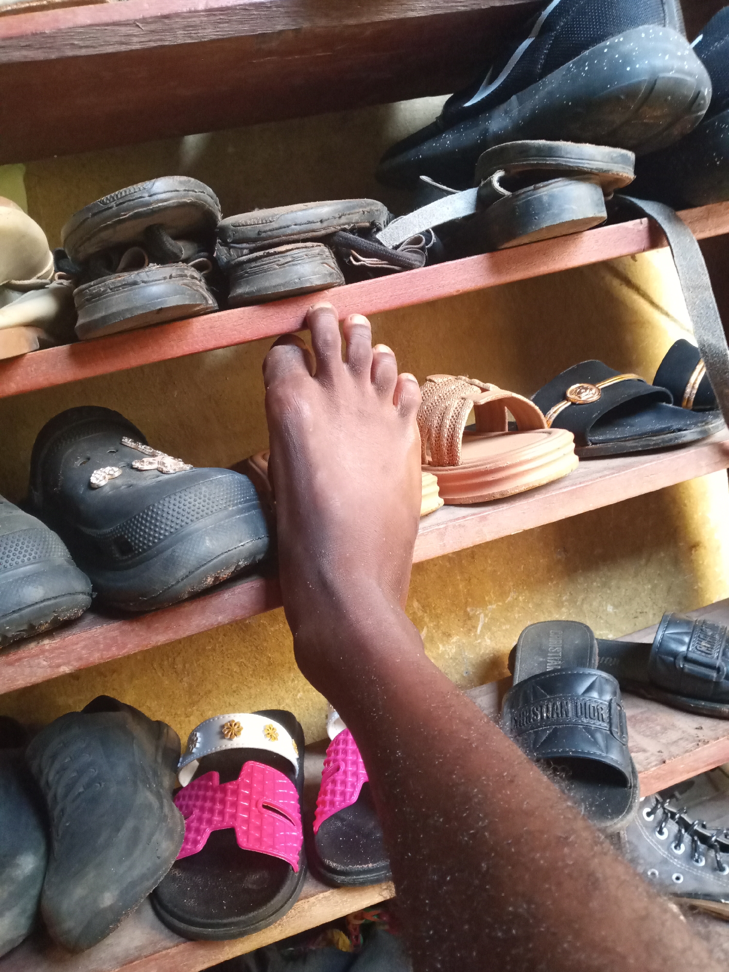 A close-up indoor shot shows a dark-skinned bare foot and forearm reaching into a worn, wooden shoe rack. The rack, with multiple shelves, is filled with a variety of shoes and sandals, arranged in a somewhat cluttered manner. The background wall appears to be a textured, light-colored surface, possibly painted or wallpapered.