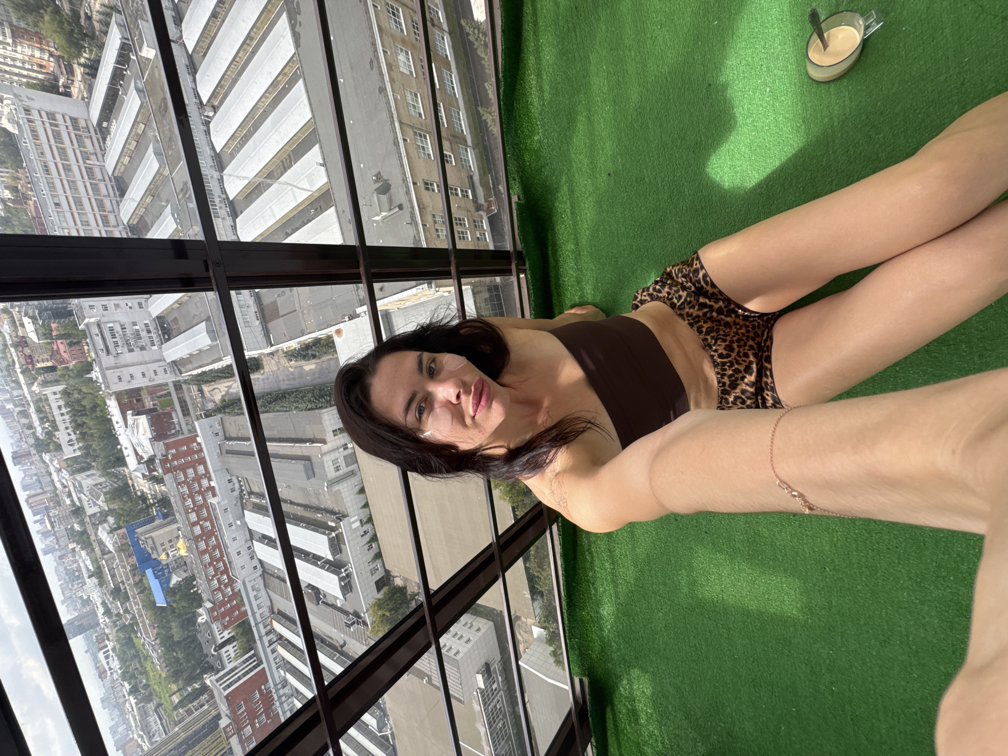 A woman with dark hair, wearing a brown tube top and leopard print shorts, sits on a green carpeted floor. She has eye patches under her eyes and is holding up her arm, taking a selfie. Behind her, a window looks out onto a city skyline with numerous buildings and trees. The scene appears to be on a balcony or enclosed outdoor space with a view of Perm, Russia. A cup of coffee with a spoon is visible on the green floor to her right.