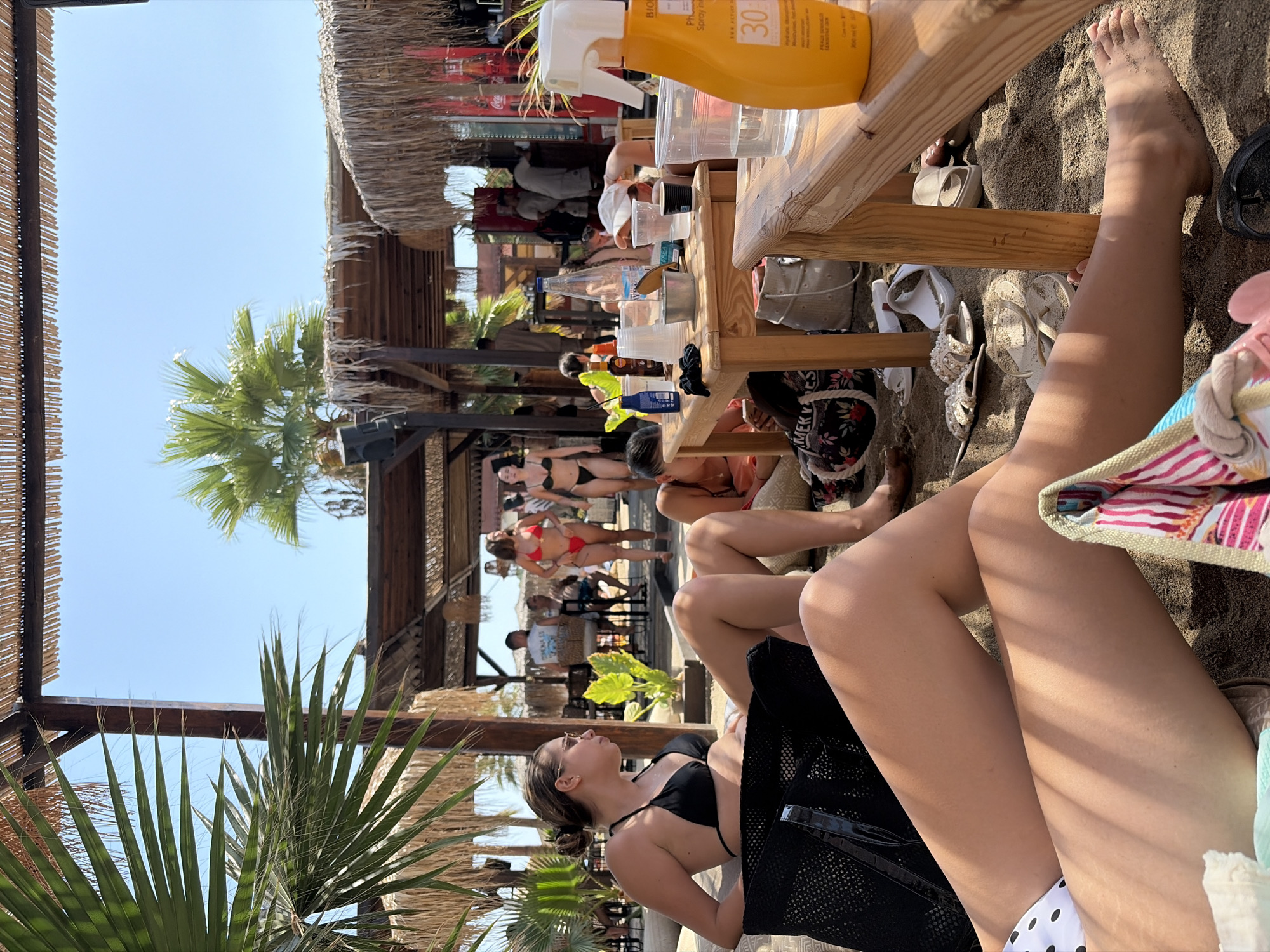 The image depicts a sunny outdoor scene at a beach bar in Kassandra, Greece. In the foreground, two women are seated on the sand, partially visible and dressed in swimwear. One woman wears a black bikini and sunglasses, while the other shows a portion of a white and black polka dot bikini bottom. Further into the scene, several other individuals are visible. Two women in bikinis walk towards the right side of the frame, one in red and the other in black. A group of people are gathered around a wooden table laden with drinks and cups, suggesting a social gathering. The environment is characterized by sandy ground, thatched roofing overhead, palm trees, and clear blue sky. The overall atmosphere appears relaxed and leisurely, typical of a beachside establishment.
