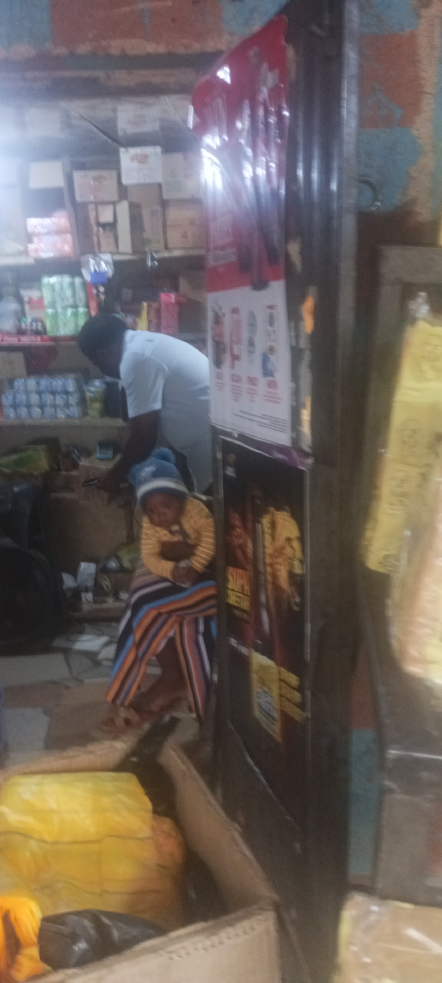 The photograph depicts a scene inside what appears to be a small store in Kuru Dungus, Nigeria. A man in a white t-shirt is reaching toward shelves stocked with various goods, including canned items and boxes. In the foreground, a child wearing a striped shirt and hat sits on what looks like a makeshift chair covered with a striped cloth. Advertising posters are affixed to the nearby structure, and boxes are visible in the foreground. The lighting is dim, and the overall impression is that of a cramped, cluttered space.