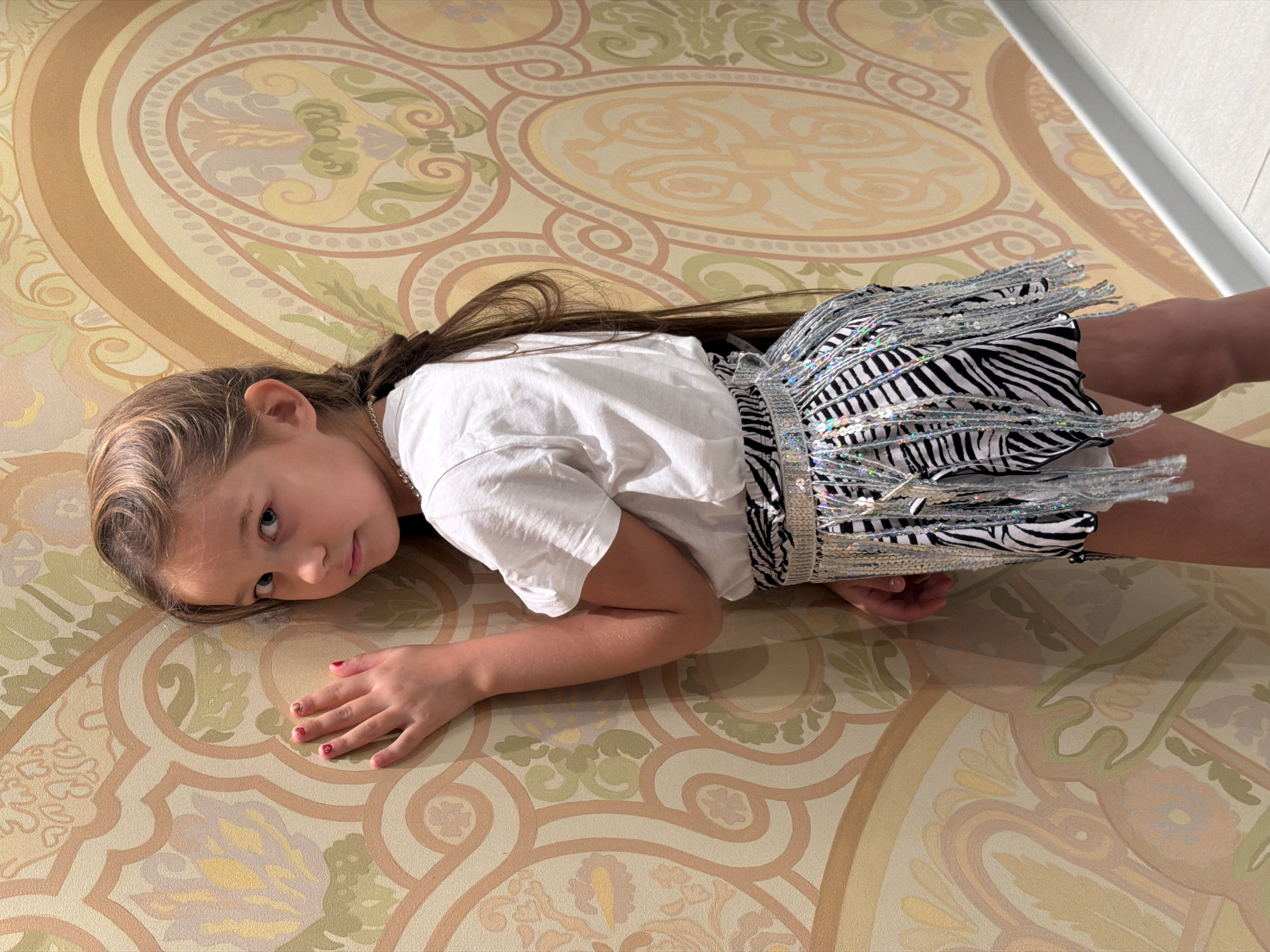 A young girl is lying on her stomach on a patterned floor. She has long, brown hair pulled back in a partial ponytail. She is wearing a white t-shirt and a black and white zebra-striped skirt with silver fringed trim. The floor has a repeating design of circular patterns and floral motifs in shades of beige, brown, and green. The setting appears to be indoors. The city of Krasnoyarsk and its location in Russia are not discernible from the provided image.