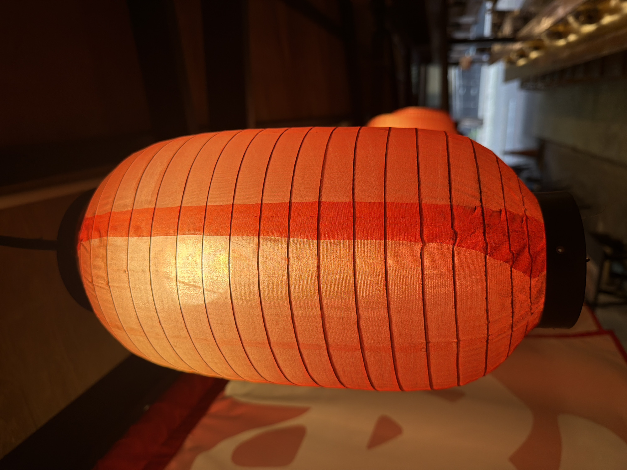 A large, elongated orange lantern hangs prominently in a dimly lit indoor setting. The lantern, the primary subject, is made of a translucent fabric or paper material, stretched over a frame that creates distinct vertical segments and a subtle horizontal band of darker orange. It glows warmly from an internal light source, casting a soft, inviting illumination. Dark, cylindrical caps are visible on both ends of the lantern.
In the background, another similar orange lantern is faintly visible, out of focus, suggesting a series of these decorative lights. The surrounding environment is dark and appears to consist of architectural elements like wooden beams or structures, particularly noticeable above and behind the lanterns. Below the main lantern, a light-colored surface with abstract, darker patterns can be seen, possibly a floor covering or part of a display.
The scene conveys a warm and cozy atmosphere, indicative of an evening or low-light indoor setting. The style of the lantern is characteristic of East and Southeast Asian cultures, aligning with the specified location of Bangkok, Thailand. There are no people visible, nor is there any discernible text within the image. The media file captures a decorative and culturally resonant element in its specific setting.