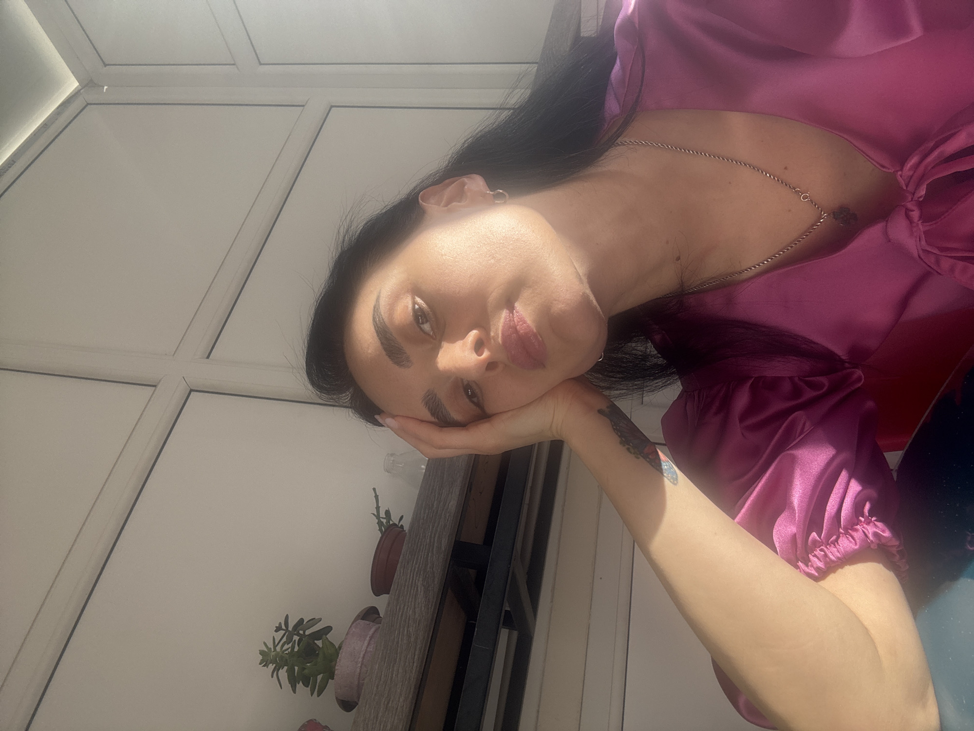 A woman with long, dark hair is resting her head on her hand, leaning to the side in an indoor setting. She is wearing a bright pink satin top. A silver necklace adorns her neck, and a tattoo is visible on her forearm. Behind her, there is a white window or wall with multiple panels, typical of modern architecture. A ledge with potted plants sits next to the window. The presence of the potted plants suggests a balcony or indoor garden area. The lighting is bright, indicating natural daylight. Given the architectural style and details, the location is likely Tbilisi, Georgia, a city known for its blend of modern and traditional aesthetics.