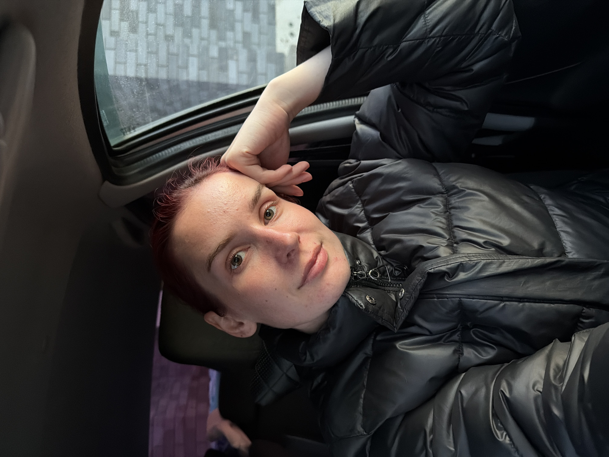 The image shows a woman inside a vehicle with an open sunroof. She is wearing a black puffer jacket. Her hair is reddish. Her right hand is near her forehead. Outside the sunroof, a blurred, urban environment is visible, possibly including buildings or structures. There is not enough detail to determine the location of the image within the city of Chaikovsky, Russia.