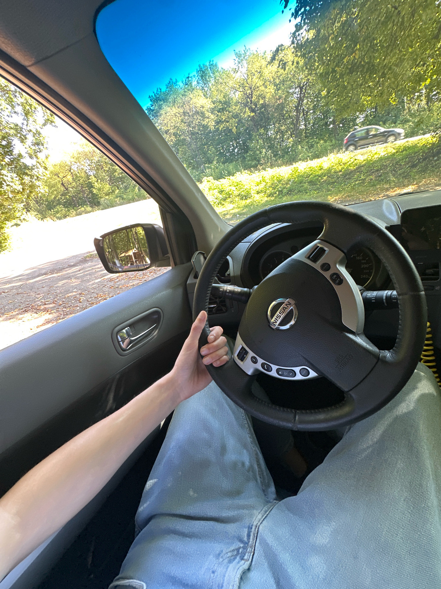 The image shows a first-person perspective from inside a car. The driver's hands are visible on the steering wheel, which has a Nissan logo. The driver is wearing light blue jeans. The car's interior, including the dashboard and door panel, is visible. Outside the car, through the windshield and side window, a sunlit, tree-lined road is seen, with another car driving on a higher incline to the right. The scene appears to be a rural or suburban setting. The image does not contain any information indicating the location is Shatava, Ukraine.