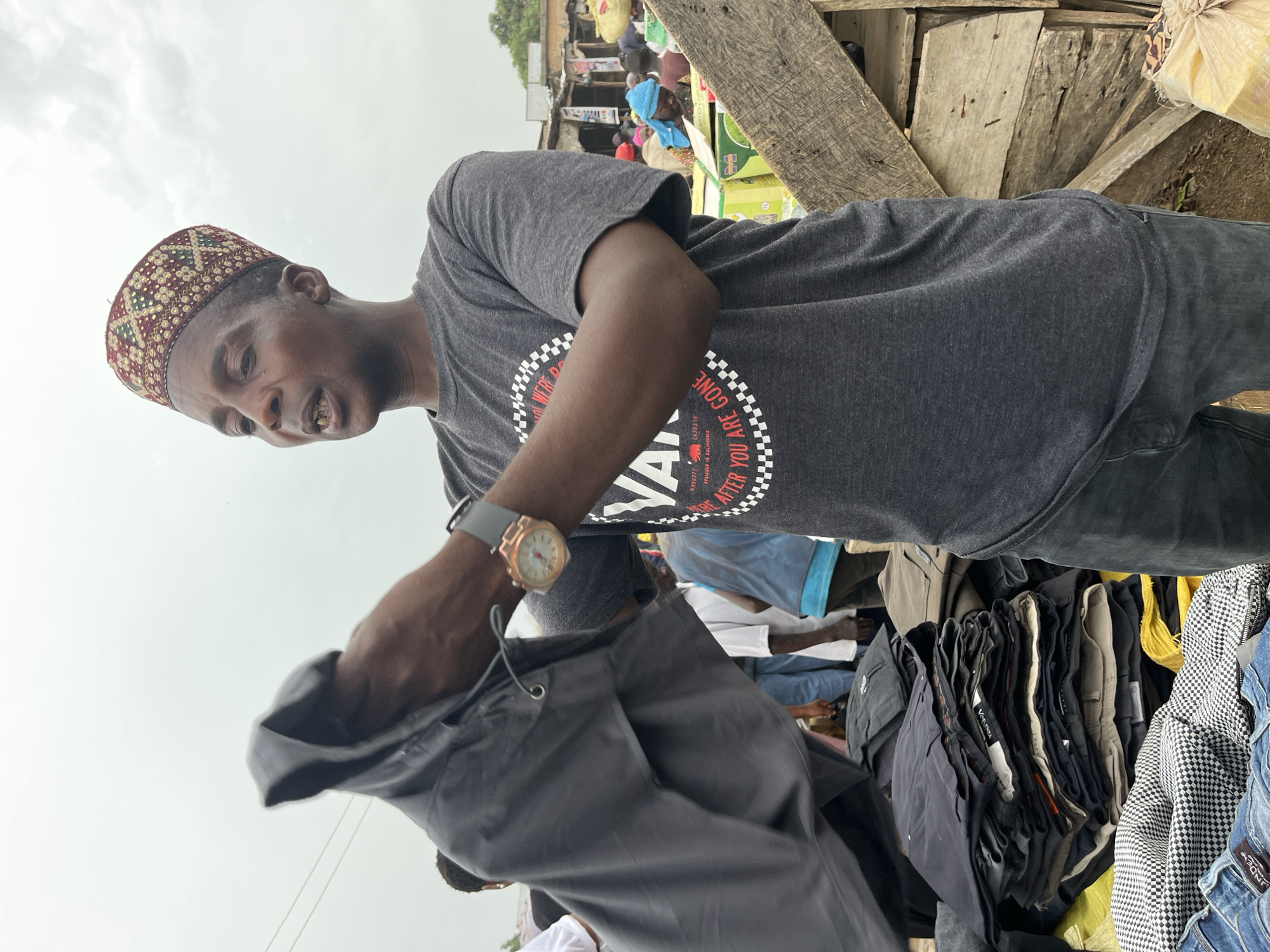 A man in Gura Top, Nigeria stands in what appears to be an open-air market. He wears a patterned cap, a grey t-shirt with a circular graphic design, and a gold wristwatch with a grey band. He is holding up a grey pair of pants, possibly inspecting it, near a pile of other clothing items. The market scene is cluttered with various objects, including wooden structures and other people in the background.