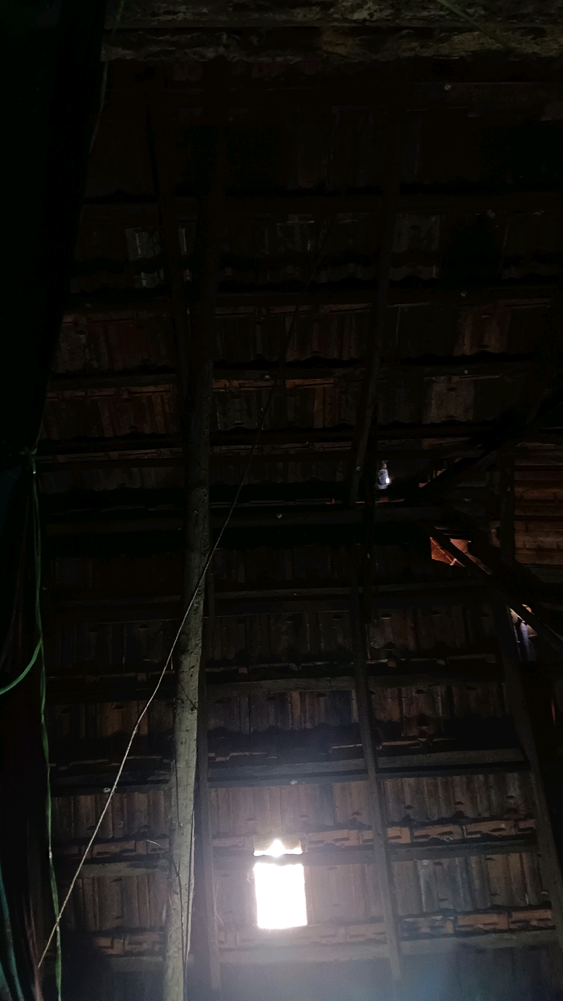The image shows the interior of a dimly lit, rustic structure, likely a barn or an old warehouse, in Punchavayal, India. The ceiling is composed of wooden beams and reddish-brown tiles, suggesting an older construction. A large, vertical wooden post runs through the center of the frame, secured by ropes and wires. A bright rectangular window with diffuse light emanates from the lower center of the image, providing the primary light source. A small, circular light bulb is visible in the upper right portion of the ceiling, emitting a faint glow. The overall atmosphere is one of disuse and age, with evident wear and tear on the wooden structures.