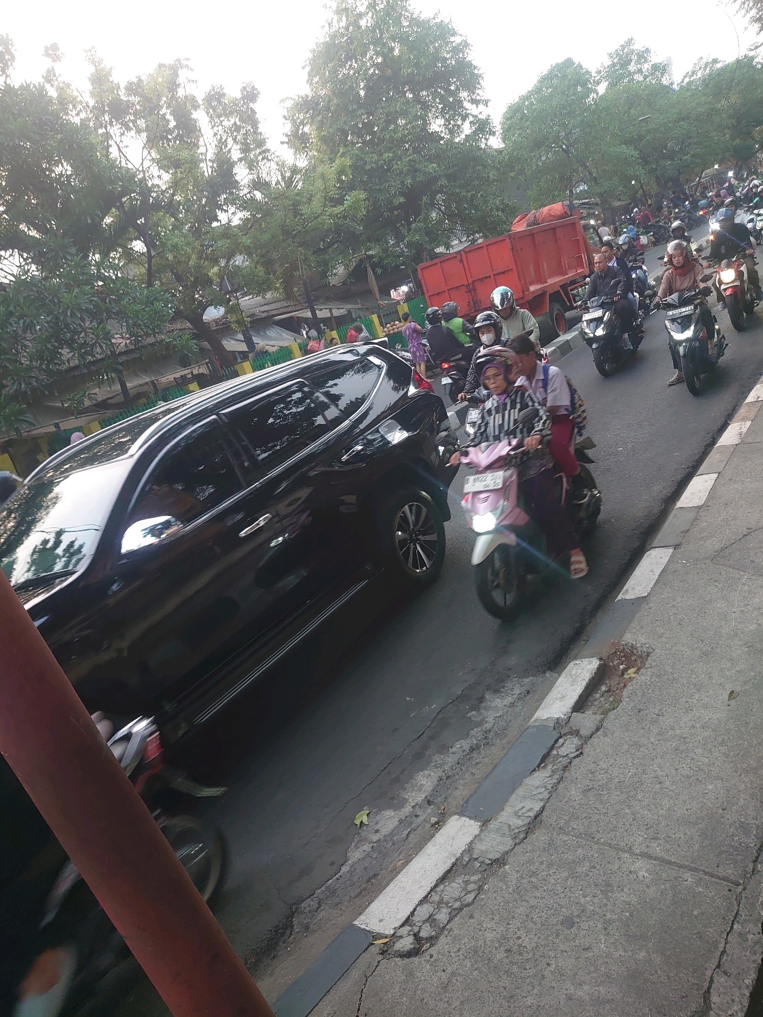 A traffic scene is captured from a slightly elevated perspective, showing a busy street in South Jakarta, Indonesia. The road is congested with various vehicles, predominantly motorcycles. In the foreground, a black SUV is parked or moving slowly on the left side of the frame. To the right, numerous motorcycles navigate the road. One pink scooter in the middle ground carries two individuals, a person driving and another passenger behind them. Several other motorcycles are visible further down the street, with riders wearing helmets. A bright red truck with a tarp-covered load is positioned in the background. Trees line the street, and buildings with awnings are partially visible behind the traffic. The lighting suggests it is either early morning or late afternoon, with the sun casting a bright glow and creating shadows. The road surface appears weathered, with visible cracks and a distinct curb on the right side.