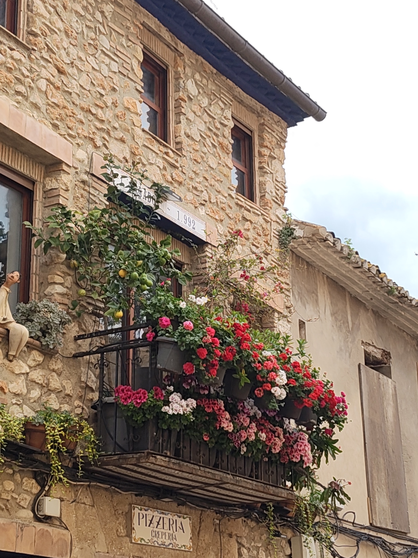 A medium shot captures the facade of a building in Dénia, Spain. The building is constructed with tan-colored stone. A balcony is decorated with a variety of flowering plants, predominantly red, white, and pink. A lemon tree grows on the balcony, bearing lemons. To the left of the balcony sits a statue of a person perched on the ledge. Above the balcony is a sign that reads "1992." Beneath the balcony is a sign that reads "Pizzeria Creperia." A separate building adjoins to the right.