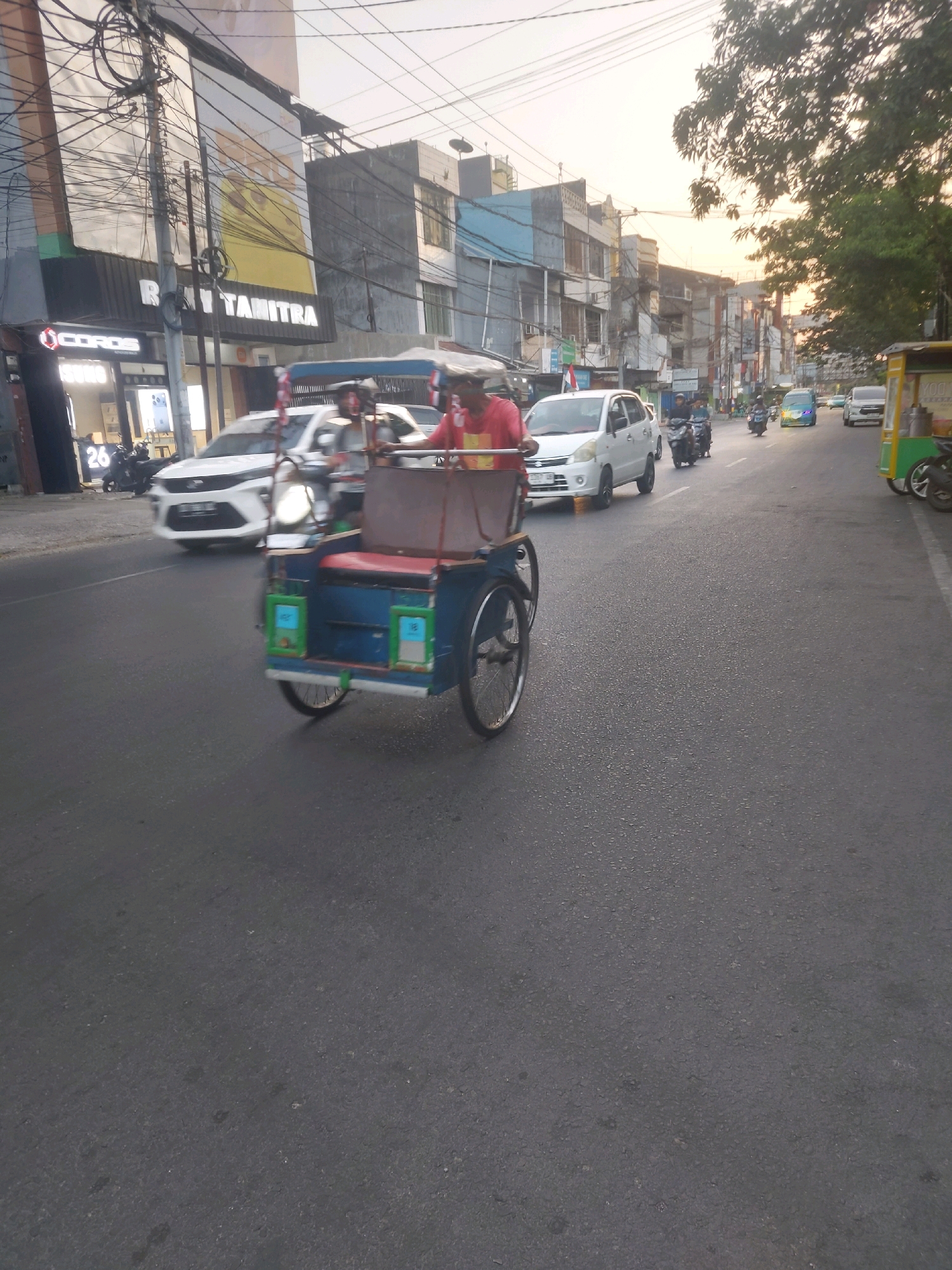 A street scene in Makassar, Indonesia, captures a bright, overcast day with a mix of buildings and vehicles. In the foreground, a three-wheeled pedicab, painted blue with red seating and green accents, is in motion, its wheels blurred from movement. A person wearing a red shirt is visible in the pedicab. The street is paved and shows signs of traffic.
Several cars and motorcycles are also present on the road. A white hatchback is seen to the left of the pedicab, and other vehicles, including a light blue van and motorcycles, are further down the street.
The background features a commercial area with various buildings. One building on the left has prominent signage, including "COROS" and "SUZUKI," along with the number "26." Another sign reads "RATU TANITRA." The architecture suggests a typical urban environment in Indonesia, with multiple stories and utility wires crisscrossing the scene. Trees with dense foliage line the right side of the street, and the sky appears bright, indicating daylight.