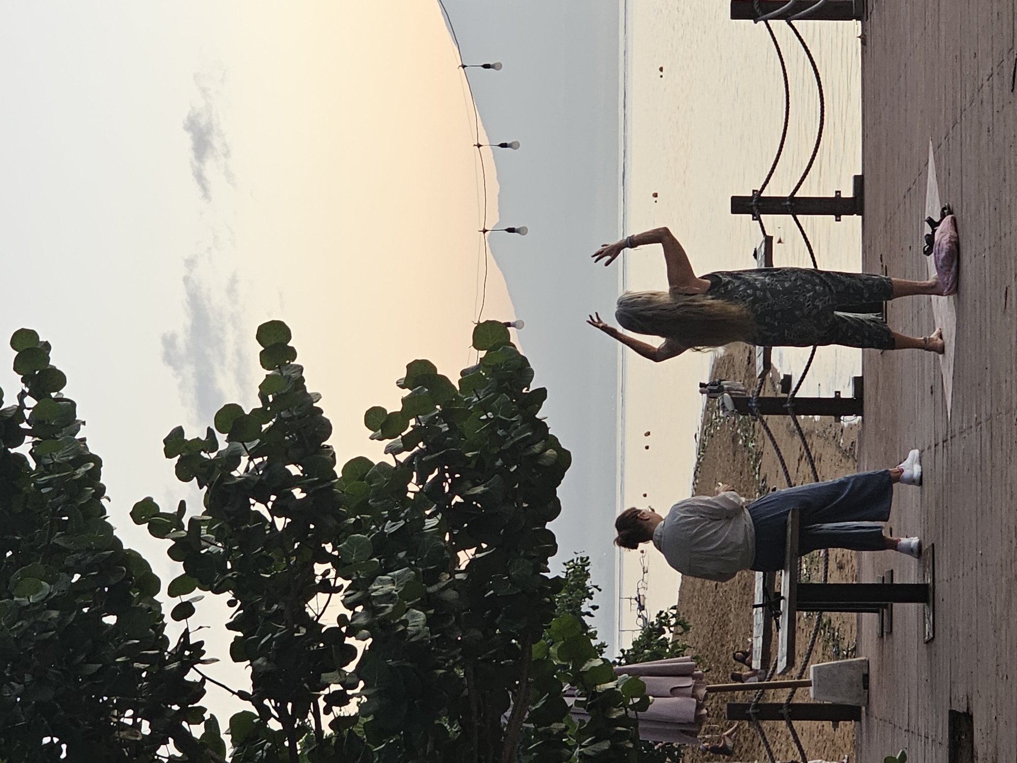 The image shows a waterfront scene in Denpasar, Indonesia, during the daytime. Two women are visible on a raised boardwalk or paved area. One woman is standing, arms raised, wearing a patterned jumpsuit and barefoot. The other woman is seated on a metal bench, facing the other direction, wearing a grey shirt, blue pants, and white sneakers.
The boardwalk is lined with metal railings supported by vertical posts. Beyond the boardwalk is a beach, with visible figures, and then the ocean. A string of lights is suspended in the background. The sky is overcast, with a hint of sunset colors. A large tree is on the left side of the frame.