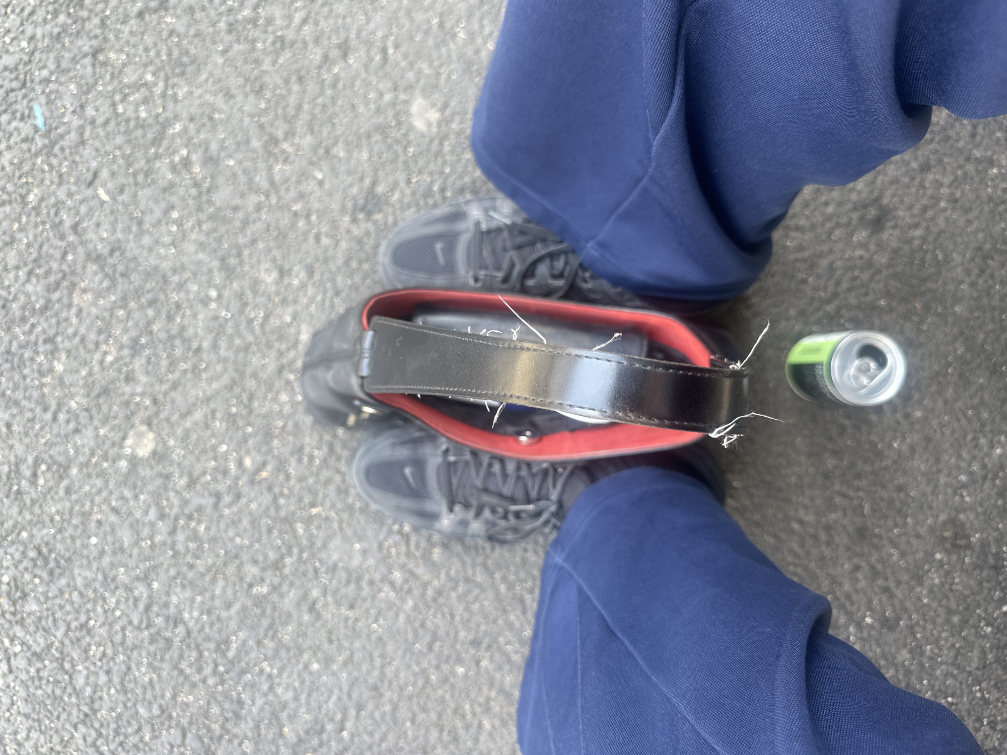 The image, taken from an elevated, first-person perspective, shows a pair of grey athletic shoes on a grey asphalt surface. The individual wearing the shoes is also wearing dark blue pants. Positioned between the feet and partially overlapping the shoes is a black bag with a red interior. A cylindrical aluminum can, green and silver in color, lies on the right side of the image, close to the bag. The asphalt surface appears to be typical of a paved area and the location might be in a city but is not necessarily identifiable as Tunbridge Wells.
