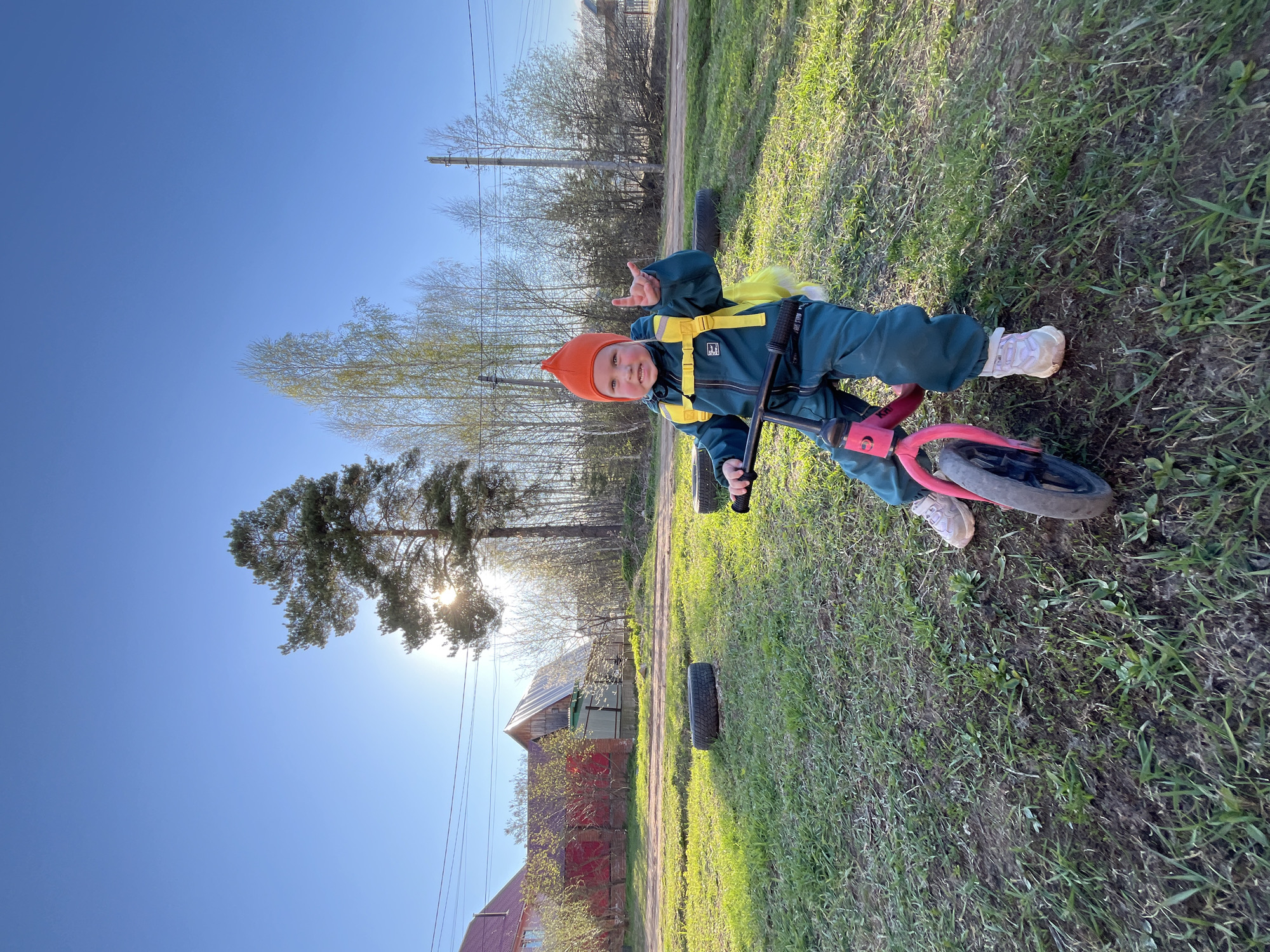 A young child is on a pink balance bicycle, giving a "hang loose" gesture with their right hand. The child is wearing a green jumpsuit with yellow straps and an orange cap. The photograph is taken outside on a grassy area. In the background is a wooden house with a red fence, a line of trees, and a clear blue sky with the sun shining through the branches of a tall tree. Two black tires are on the ground as obstacles. It's a sunny day. The scene appears to be in a rural area, possibly Chaikovsky, Russia.