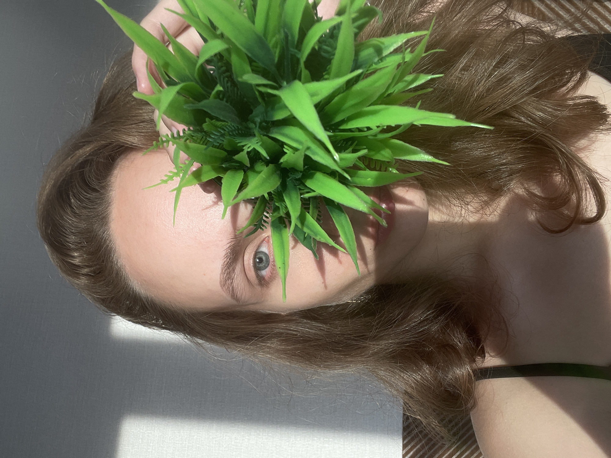 A person lies on a textured surface, possibly a bed, with their head tilted to the side. They are holding a cluster of artificial green foliage over their face, obscuring their nose and mouth. One eye is visible, a striking blue-grey, looking directly at the camera. The person has long, wavy brown hair that falls around their head and shoulders. Sunlight casts a distinct shadow across the lower left portion of the image, suggesting it is daytime and potentially near a window. The background is a soft, neutral tone with subtle stripes in the lower right corner. The scene appears to be indoors, likely in a bedroom or a studio. There is no visible text. The overall mood is enigmatic and artistic, with the play of light and shadow, and the partially obscured face.