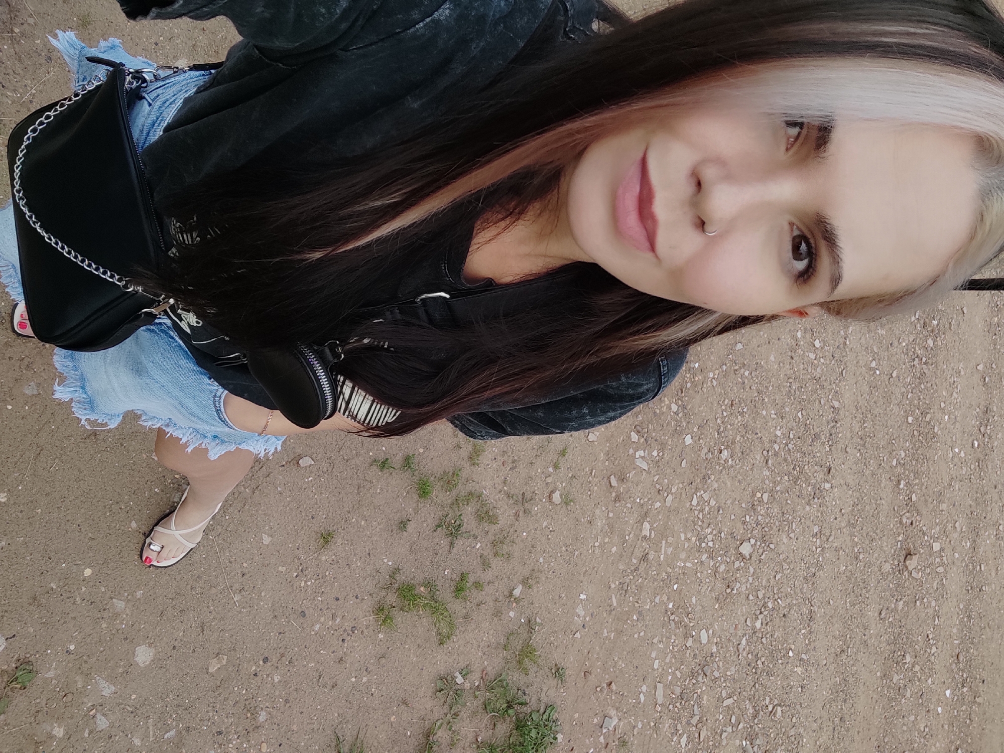 This is a high-angle, medium shot of a young woman standing on a dirt path. She is wearing a black jacket, a light blue denim skirt with a frayed hem, and white strappy sandals. A black purse with a silver chain strap is slung over her shoulder. The woman has long, dark hair with blonde and pink highlights. She is looking at the camera with a slight smile. Her facial features include a nose ring and light makeup. The ground is composed of dirt, small stones, and sparse green grass. The overall lighting suggests it is daytime, possibly overcast or late afternoon. The background is out of focus, indicating the subject is the primary focus of the image. The setting appears to be an outdoor, somewhat rural or unpaved area, consistent with a location like Ольховка, Russia. There is no visible text in the image.