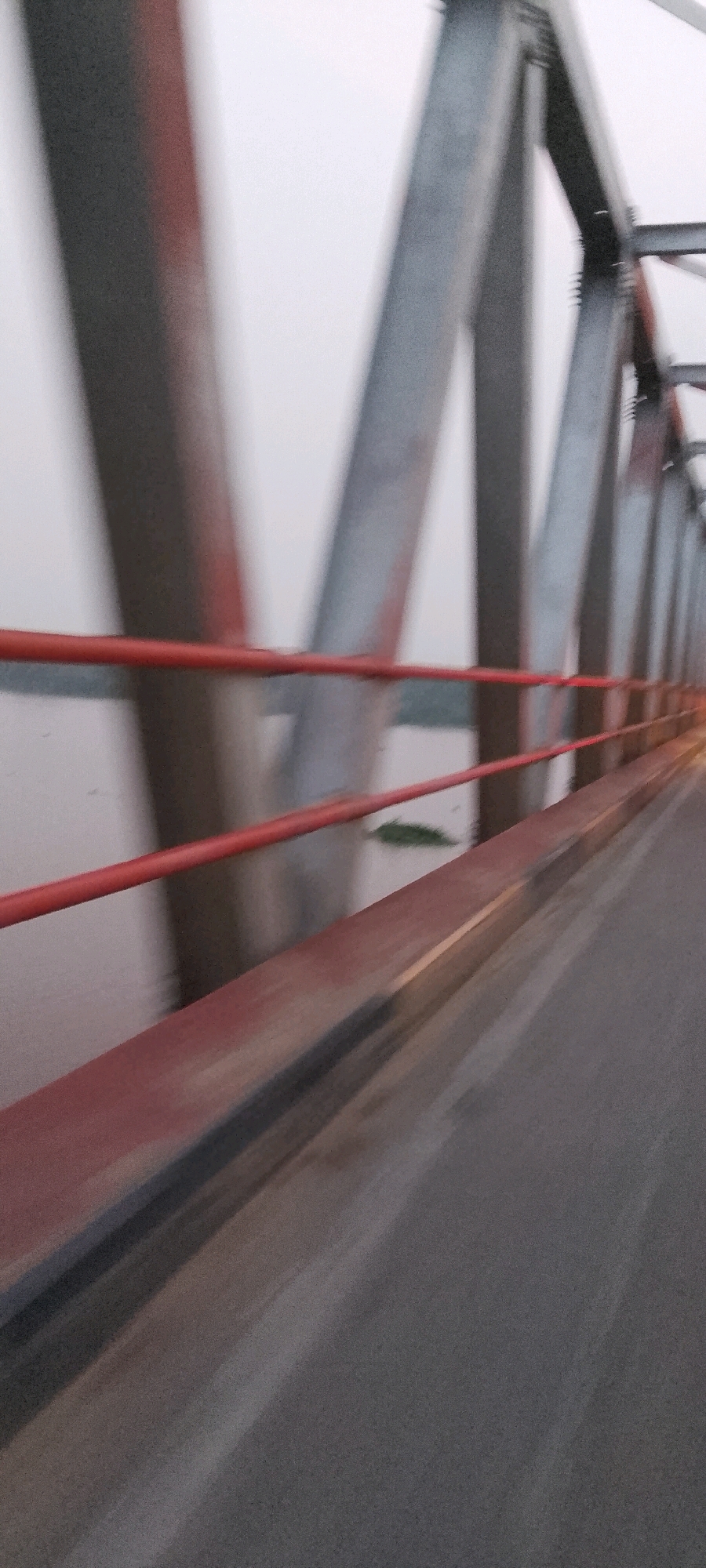 A blurry, low-angle, outdoor, mid-day shot shows a roadway on a bridge in Jumrah, Indonesia. The bridge is made of a grey metal truss structure with red railings. The roadway appears to be asphalt. A body of water is visible below the bridge, with a small patch of green vegetation. The sky is overcast and grey. The motion blur suggests the photo was taken from a moving vehicle. There are no people visible in the image.