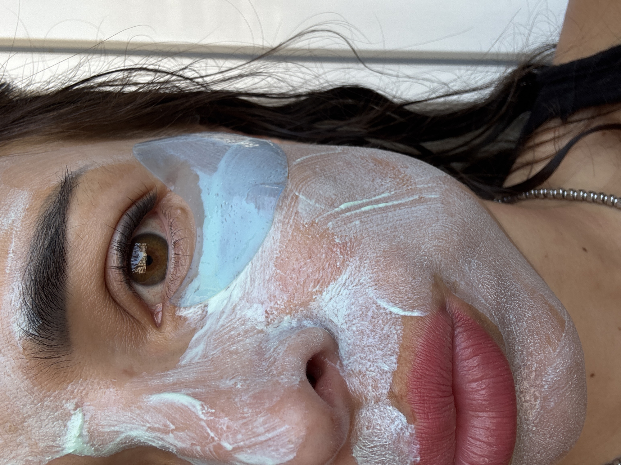 A close-up, right-facing, shoulder-up shot shows a person's face, partially covered in a white face mask and blue under-eye patches. The person has brown eyes, dark eyebrows, and dark hair that falls over their shoulder. Their lips are slightly parted and appear to be colored with lipstick. The background is blurred and appears to be indoors, with a white wall or partition visible. The lighting is natural and seems to be coming from the left. No city or location information is available.