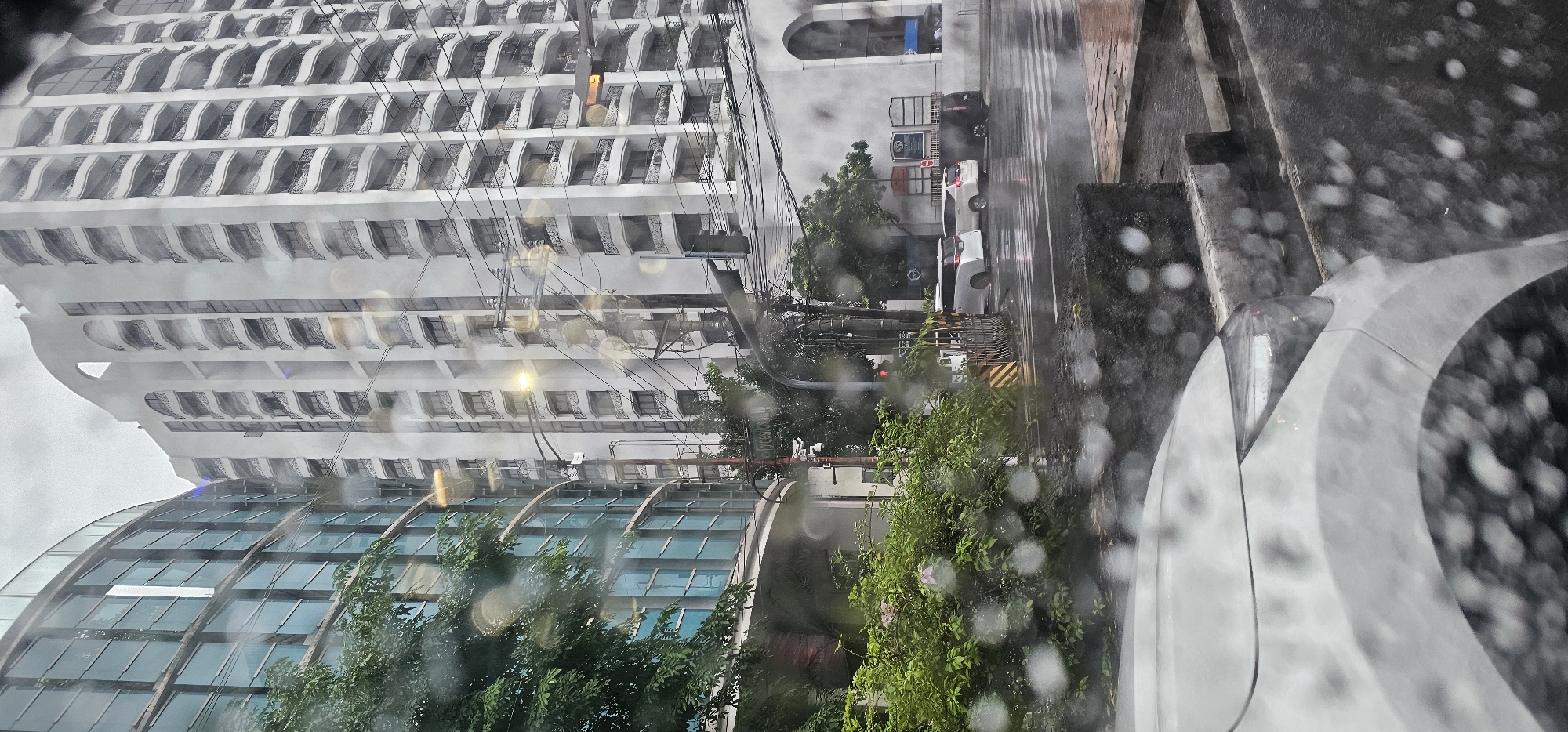 The image depicts a street scene in Makati, Philippines, during what appears to be a rainy day, as evidenced by the water droplets on the foreground window and the wet road. The view is from inside a vehicle, looking out.
In the background, a tall, white, modern building with a repeating pattern of balconies and arched window frames dominates the right side of the frame. To the left of this building, a curved structure with a glass facade, likely another building or part of a larger complex, is visible. Green trees and dense foliage are present in the midground.
The street shows signs of traffic, with several vehicles visible. A white minivan, a white SUV, and a dark SUV are parked or moving on the wet asphalt road. A pedestrian crossing is marked on the road. Traffic lights and utility poles with numerous wires are present throughout the scene, contributing to the urban environment. A street sign indicating "J. P. Rizal" is partially visible. The overall atmosphere is overcast and damp, characteristic of a rainy urban setting.