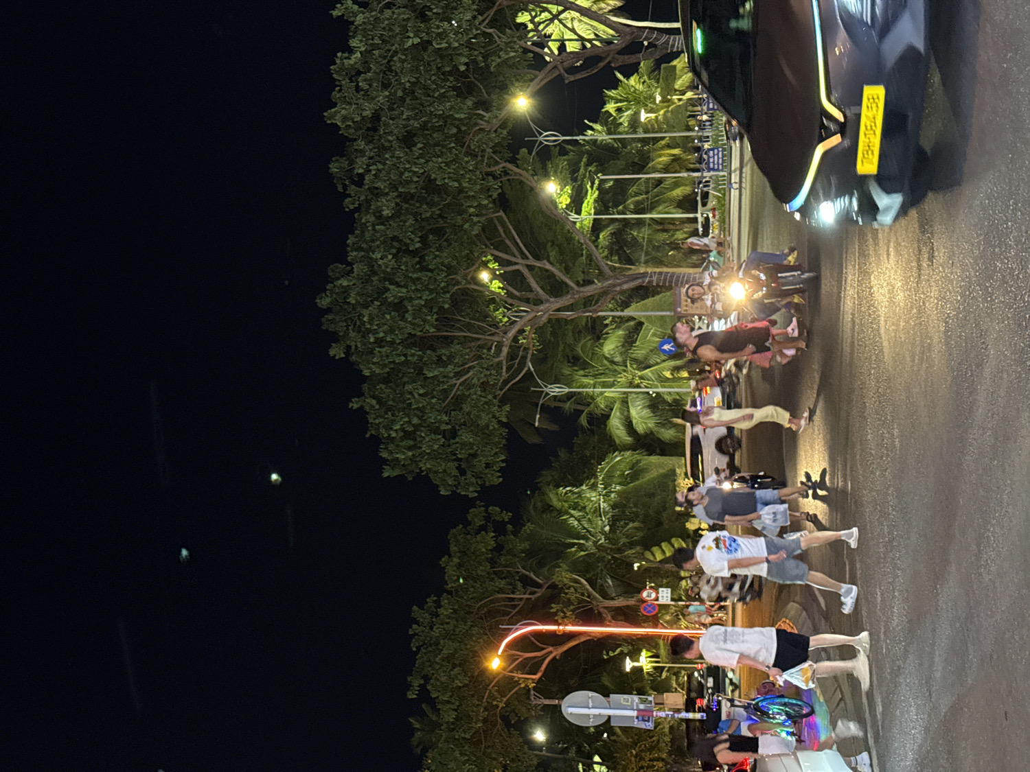 The image captures a bustling street scene at night in Nha Trang, Vietnam. The perspective looks along a road lined with numerous trees, including prominent palm trees and leafy green foliage, silhouetted against the dark sky and partially illuminated by artificial lights.
Several people are present, engaged in various activities. On the left side of the street, individuals are seen walking casually, dressed in summer attire such as shorts, t-shirts, and dresses. Further down the road, closer to the center, a group of people is gathered around motorcycles, with one person appearing to be riding a motorcycle with its headlight brightly lit. Another person is seen crossing or standing near the road.
To the right, a dark-colored car is partially visible, facing away from the viewer. Its rear lights are illuminated, and it features a distinctive bright yellow license plate with the text "BE-750-HEL".
The scene is brightly lit by multiple light sources, including streetlights, vehicle headlights and tail lights, and ambient light, creating reflections on the seemingly damp road surface. The overall impression is that of an active urban night environment, with the trees adding a tropical feel to the setting. Street signs are visible but mostly unreadable due to distance and lighting, with one blue sign displaying a white arrow.