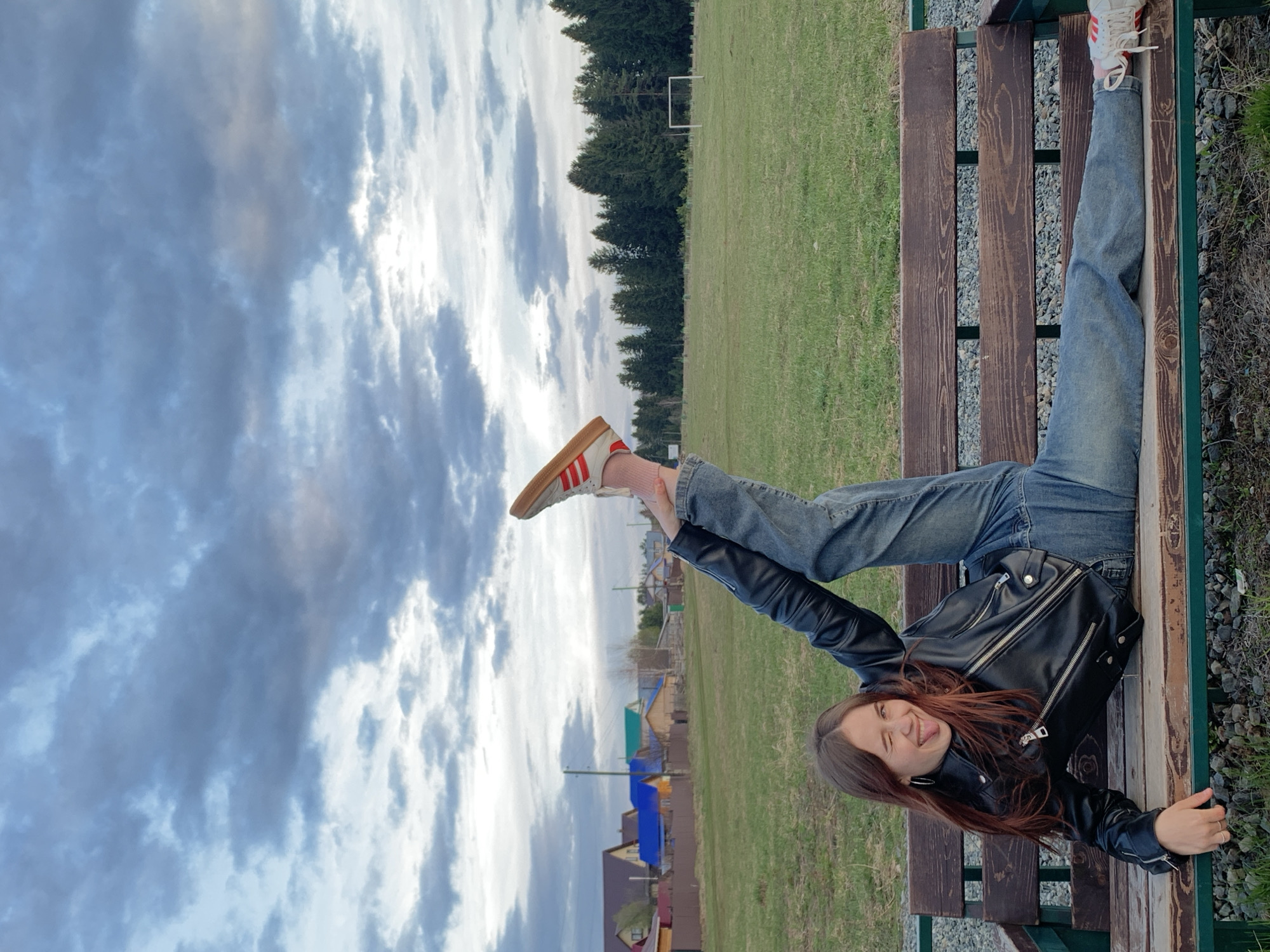 An outdoor, sideways-oriented photograph captures a young woman playfully posing on a wooden park bench. She is lying on her back, looking towards the camera with a wide smile and her tongue sticking out. Her head and upper body are slightly raised, and her legs are bent at the knees, with her feet, clad in white sneakers with red stripes and pink socks, pointing upwards. She is wearing a black leather jacket and blue jeans.
The park bench, made of dark wooden slats supported by green metal frames, sits on a surface of gravel and grass. In the background, a large green field extends, with a white football (soccer) goal visible in the distance on the left. A line of dense evergreen trees borders the field. Beyond the trees and field, several houses with varying colored roofs (blue, orange, green) can be seen, suggesting a rural or village setting, characteristic of Фоки, Russia. The sky overhead is largely overcast with soft, scattered clouds, indicating a daytime scene with diffused light. There is no visible text in the image.