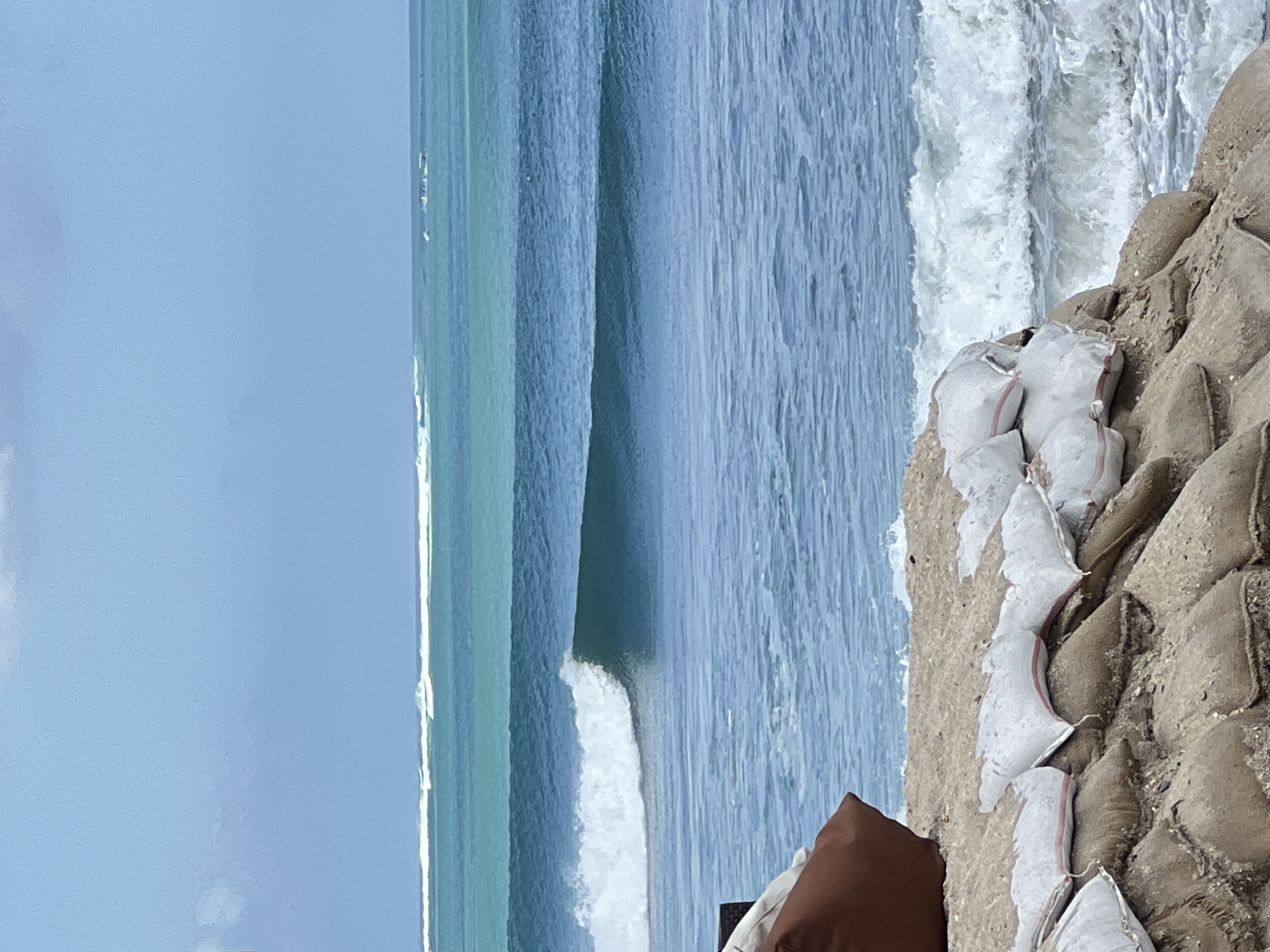 This image captures a segment of the coastline in Kuta, Indonesia, facing the ocean under a bright, partly cloudy sky. In the foreground, a sandbag barrier is constructed, composed of numerous stacked sandbags, some covered with white material and others appearing in their natural tan color. A portion of a large, brown beanbag chair is visible on the sand above the sandbags.
The middle ground of the image is dominated by the ocean. A significant wave is breaking on the left side, showing a clear curl and white foam. Further out, two distinct lines of breaking waves are visible on the horizon, suggesting multiple reef breaks or a strong swell. The water transitions in color from a deep blue closer to the shore to a vibrant turquoise further out, indicating varying depths or water clarity. A small white boat is visible in the distance on the right, on the turquoise section of the water.
The sky above is a pale blue with scattered white clouds, and the overall impression is one of a calm yet active coastal environment, likely prepared for potential coastal erosion.