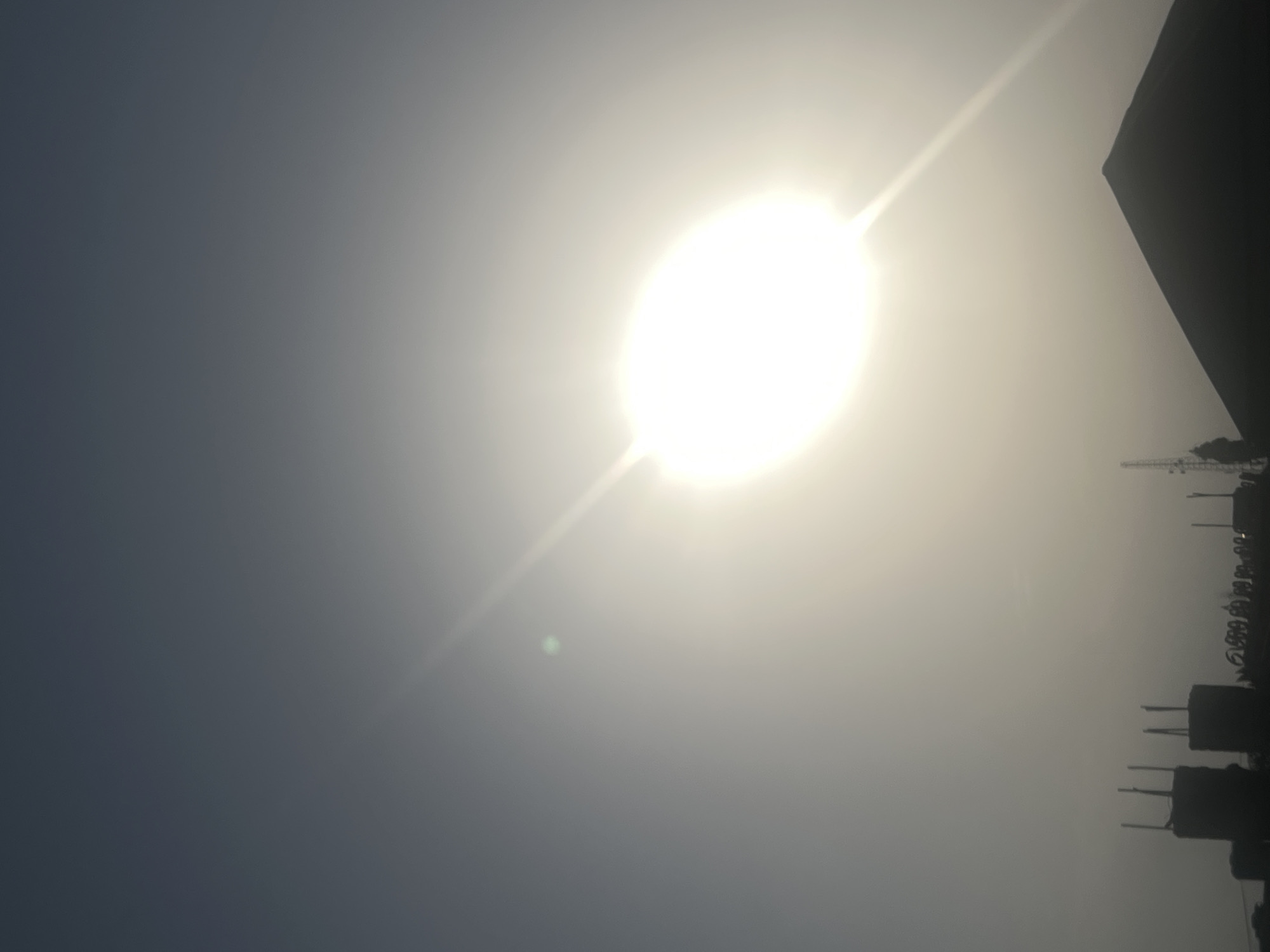 The image captures a hazy sky over Jalingo, Nigeria, with the sun prominently displayed and slightly overexposed, casting a bright, diffused light. The sun appears as a large, luminous oval, with noticeable light flares radiating outwards, particularly in the upper right quadrant. A small, greenish lens flare is also visible in the sky. Silhouetted against this bright sky are the rooftops and structures of Jalingo. In the lower portion of the image, the dark shapes of buildings are discernible, including what appear to be construction sites with exposed rebar or structural elements sticking out of the tops of some buildings. A triangular rooftop dominates the lower right section, and a cellular tower or transmission mast is visible to the left of it. The overall atmosphere is muted due to the haze and the bright sunlight.